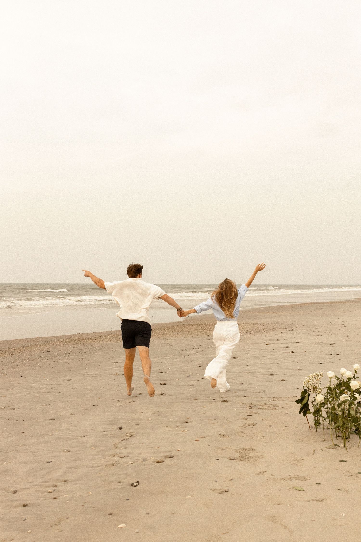 A joyful couple runs hand-in-hand along a sandy beach toward the ocean, with white flowers in the foreground.
