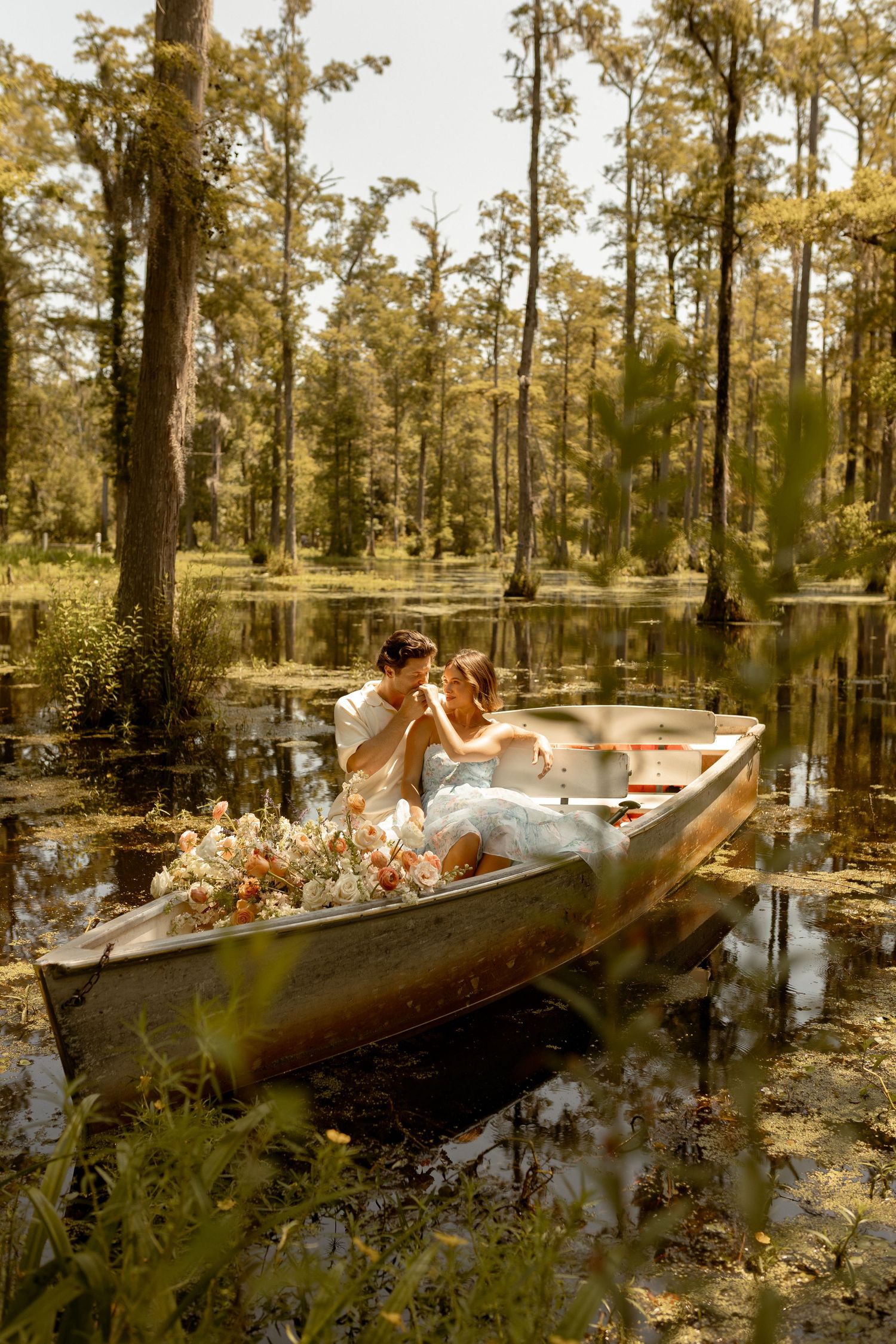 Couple kissing in a flower-filled rowboat on a serene swamp surrounded by cypress trees.