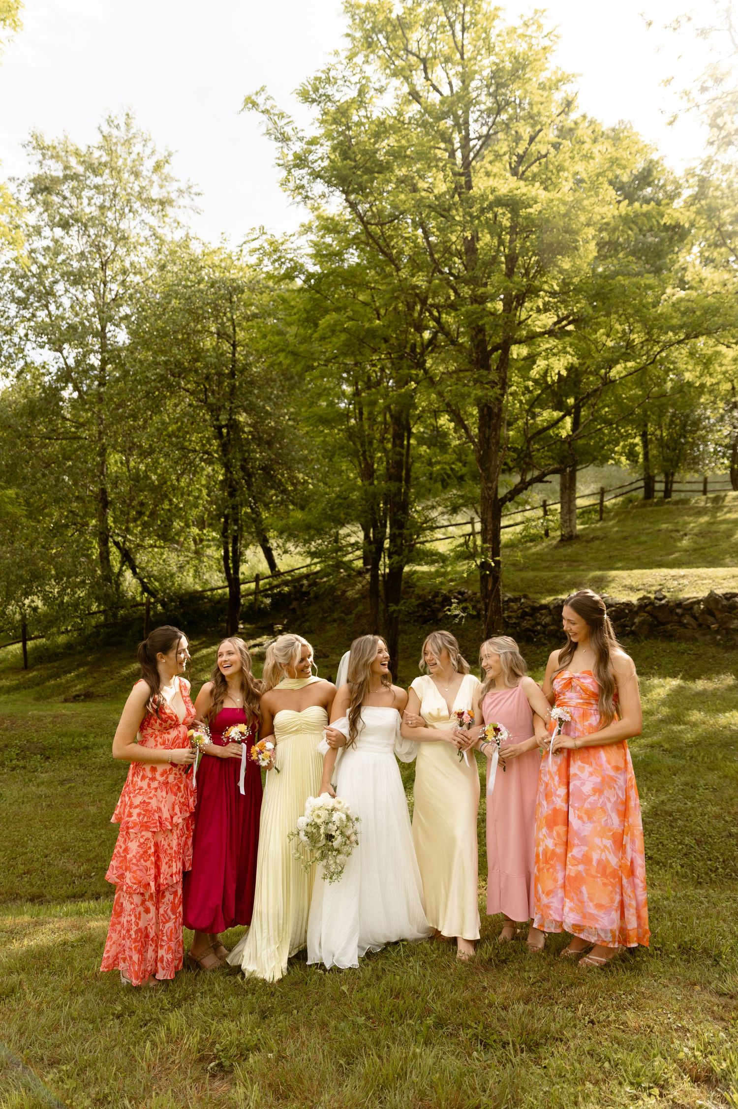 Bride in white gown stands with six bridesmaids in colorful dresses on a green hillside at an outdoor wedding.