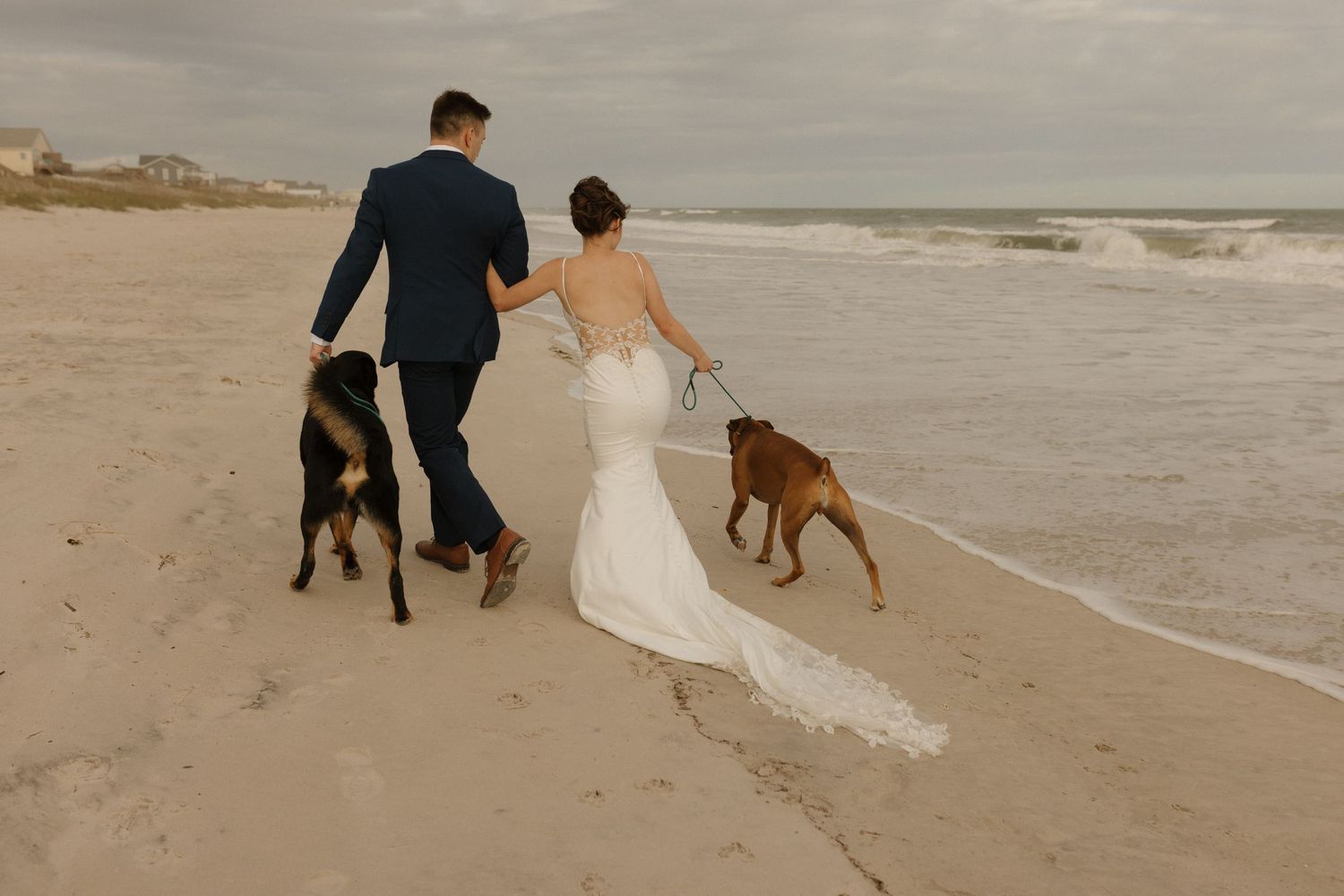 Bride and groom walking on beach with two dogs, waves in background, wedding dress train trailing on sand.