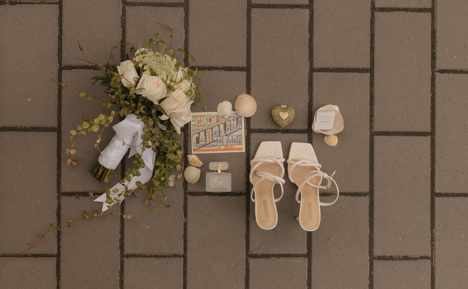 Flat lay of bridal accessories including white roses, nude heels, and wedding details on gray pavers.