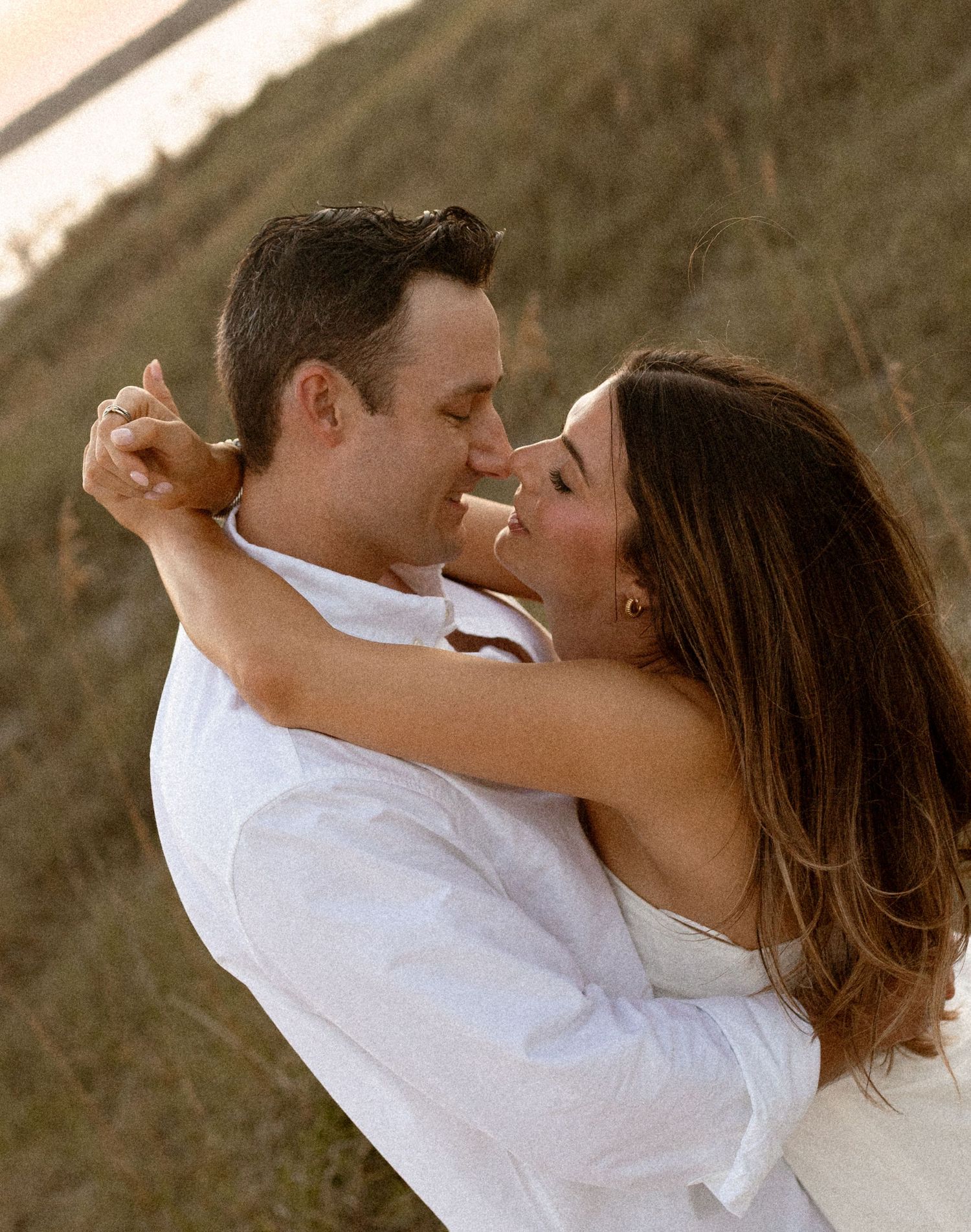 A couple in white clothing embraces and nearly kisses outdoors in a golden field during a romantic engagement photo session.