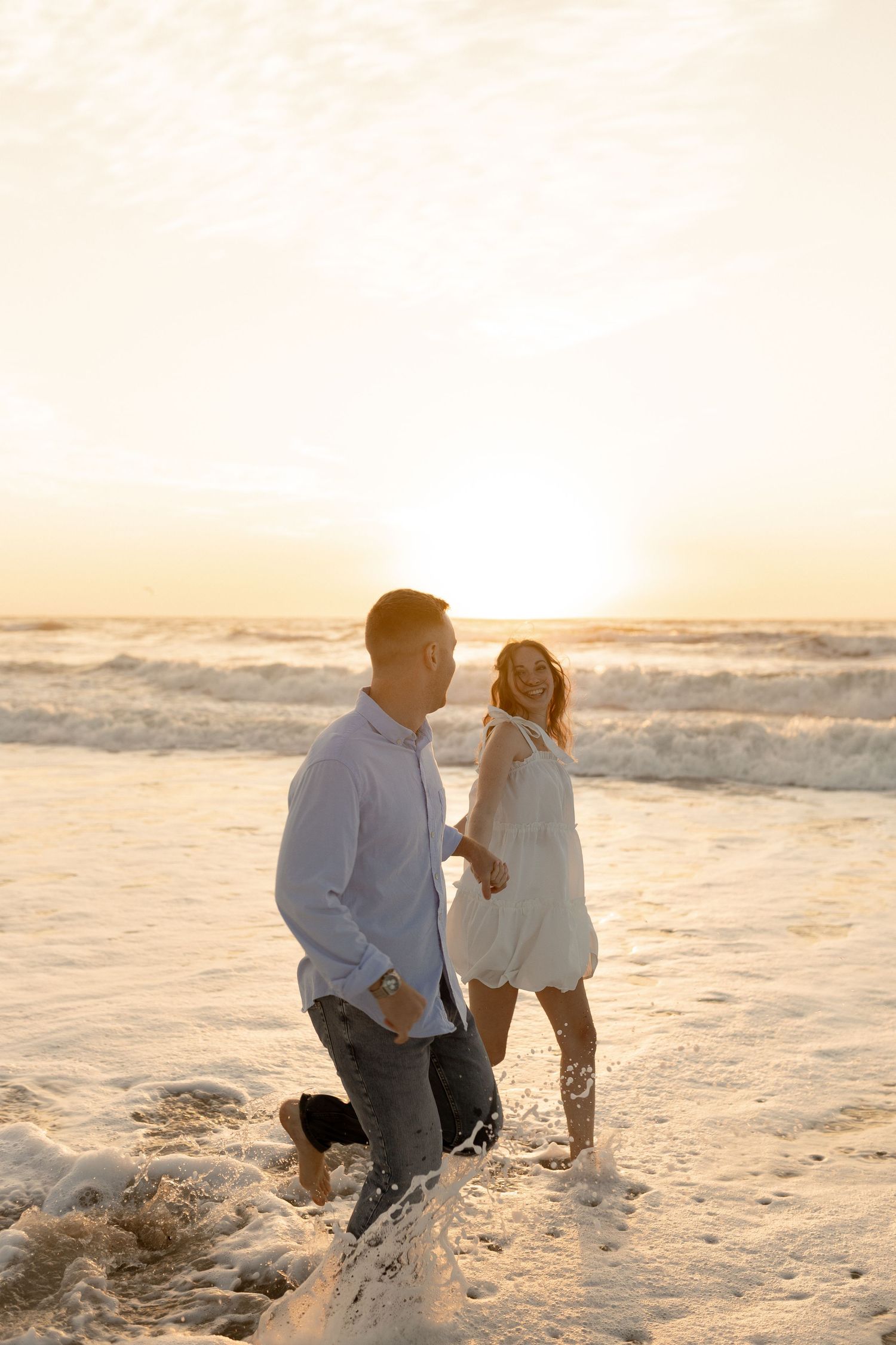 Couple walking hand in hand on a beach at sunset, waves splashing around their feet.