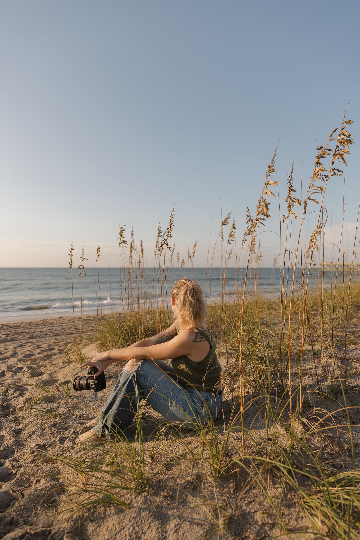 A blonde woman sits in beach grass dunes holding a camera, gazing at the ocean at golden hour.