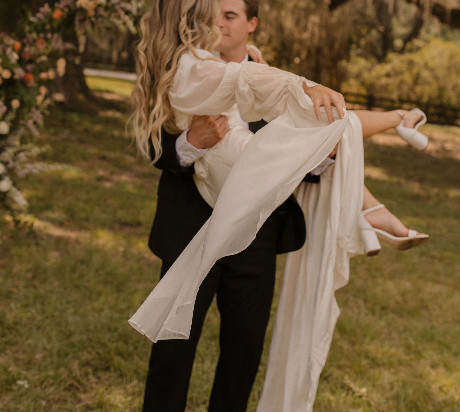 Groom in black suit carrying bride in white wedding dress outdoors.
