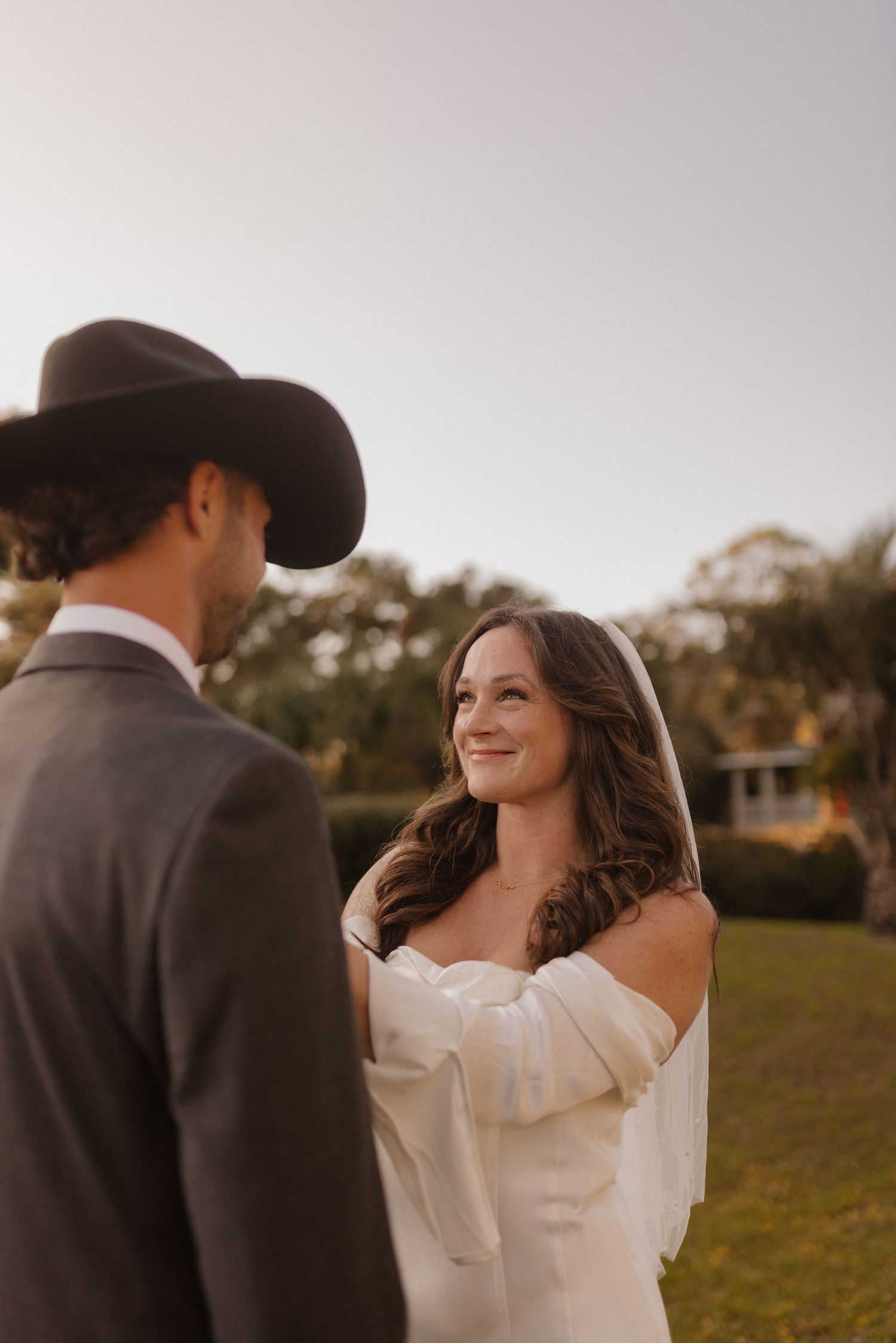 Bride in off-shoulder white gown and veil smiles at groom wearing a black cowboy hat outdoors.