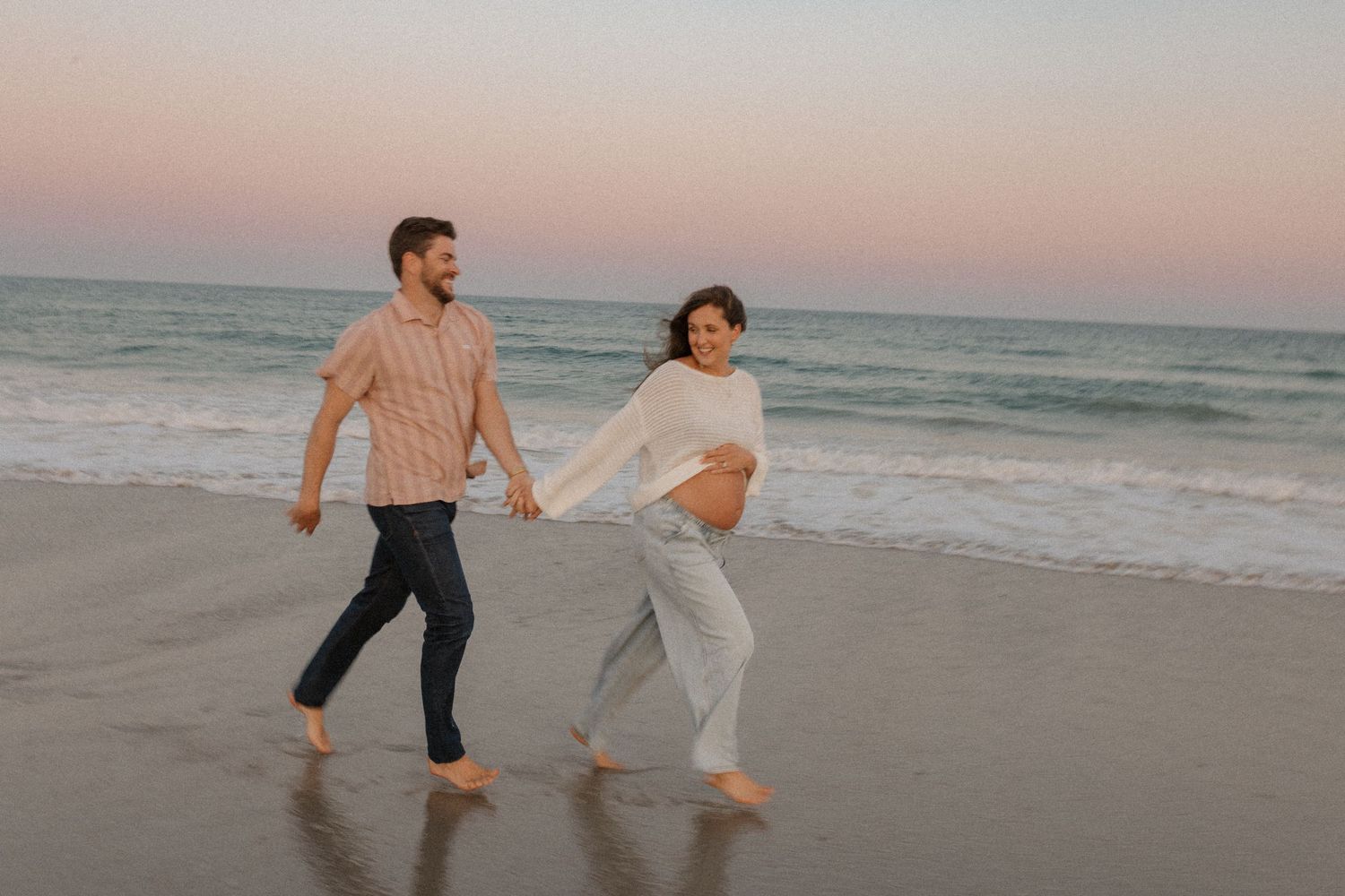 Pregnant couple walks barefoot along the shoreline at dusk, holding hands and smiling.
