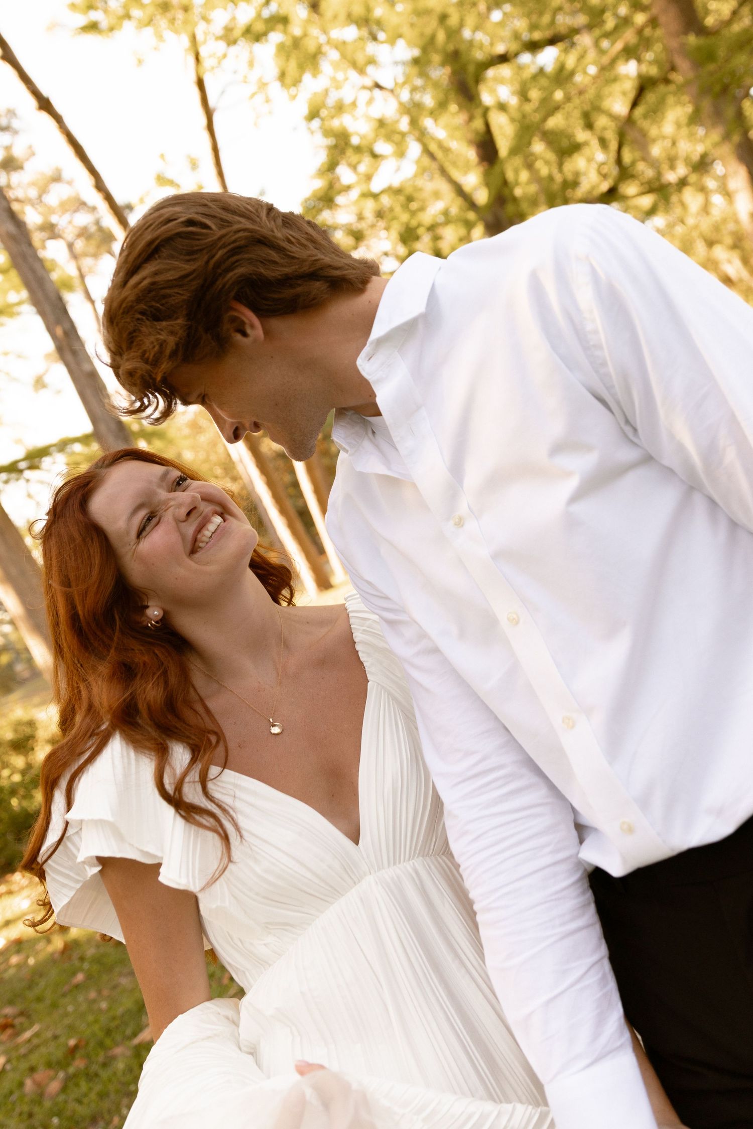 Joyful red-haired bride in white dress laughs with groom in white shirt among golden autumn trees.