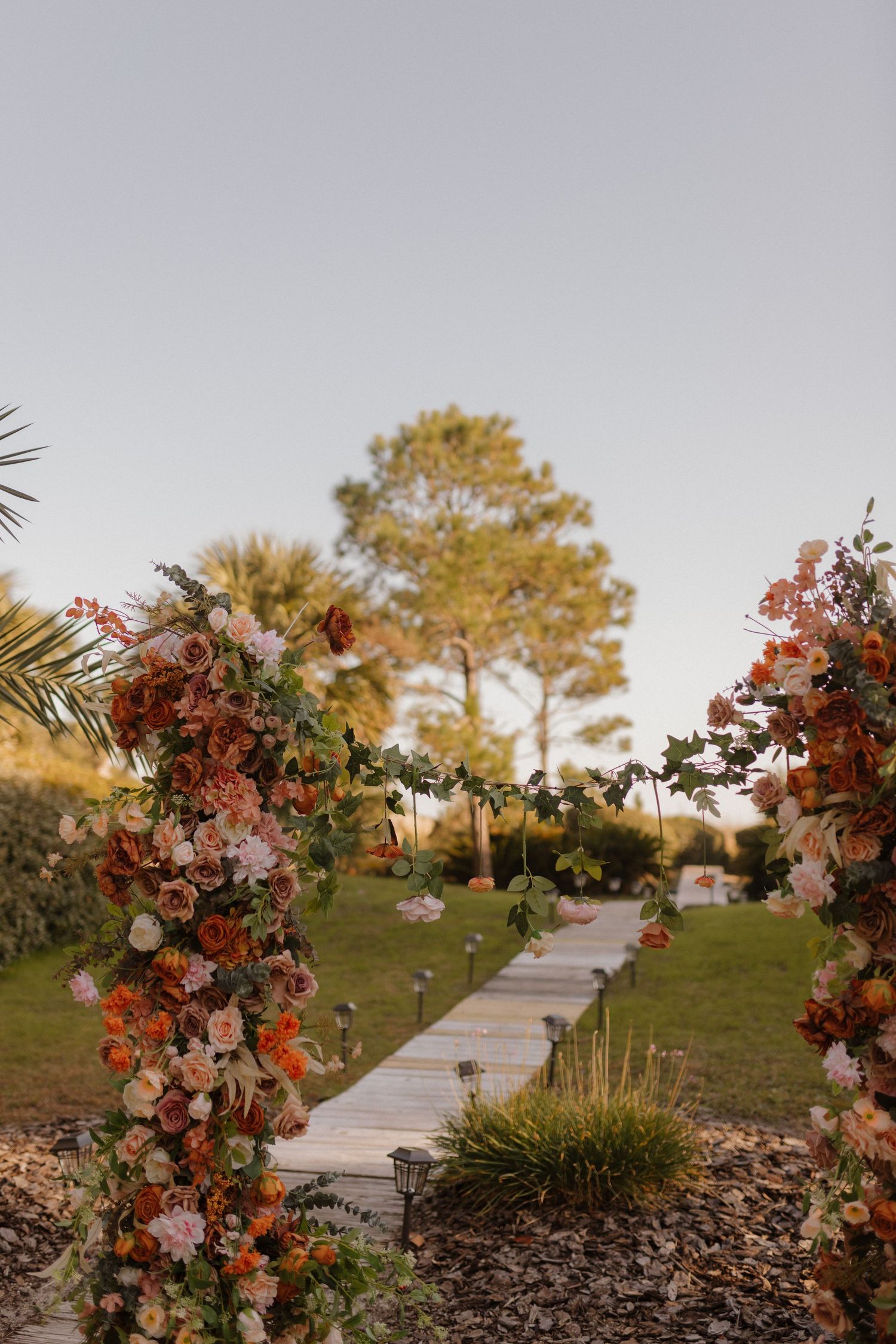 Rustic outdoor wedding aisle with floral arch columns of rust and pink roses at golden hour.