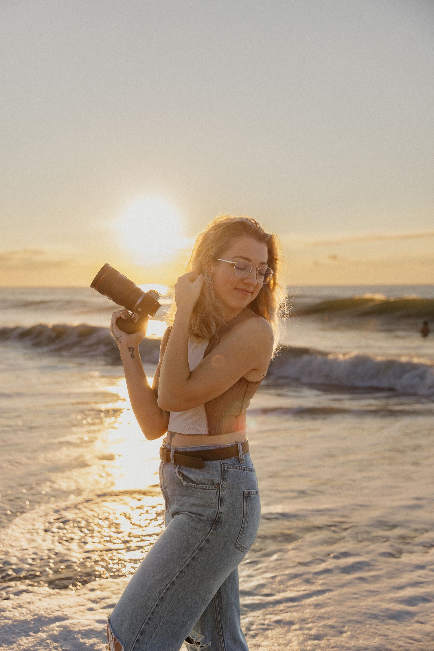 Female photographer in jeans and crop top holds camera at golden hour sunset on beach.