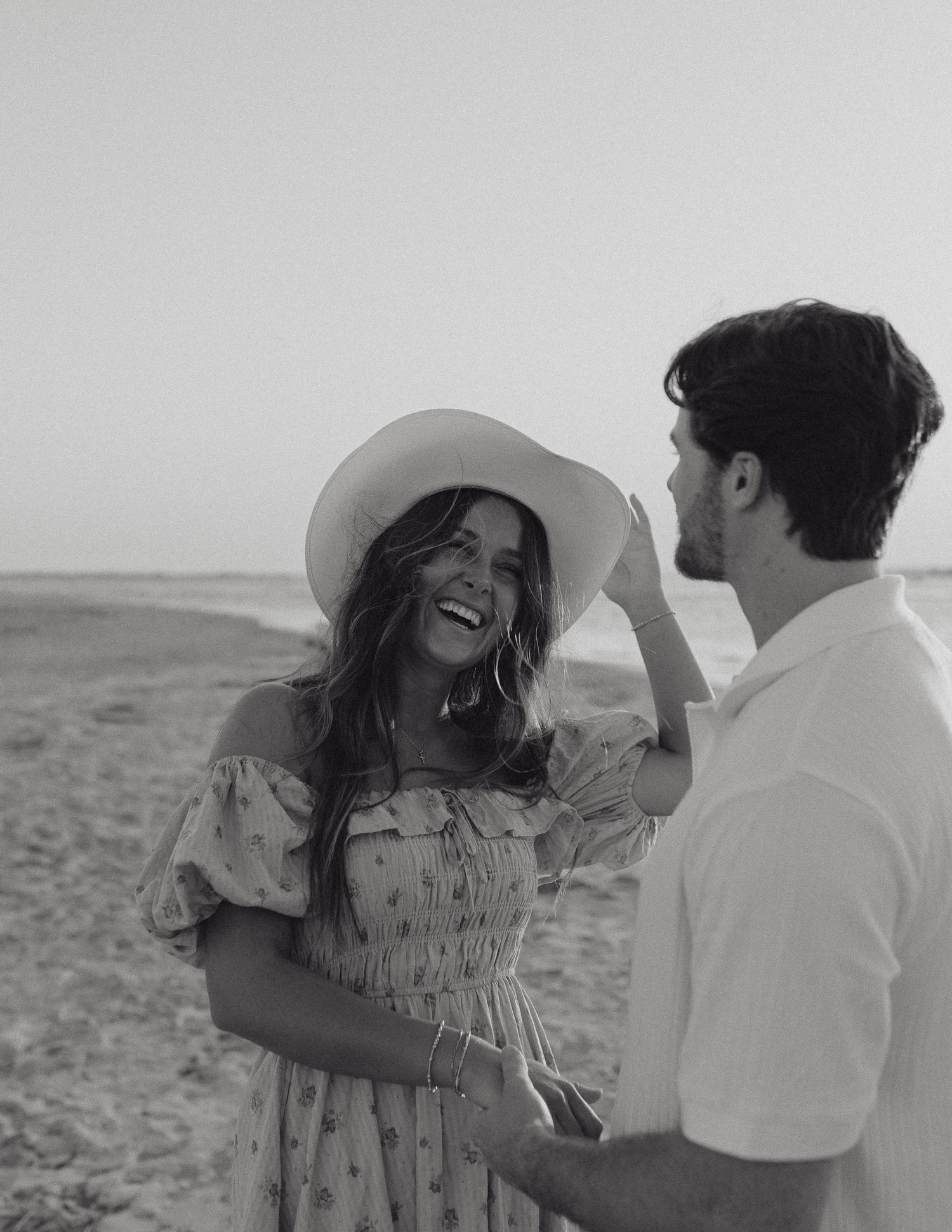 Joyful woman in floral off-shoulder dress and sun hat laughs with man on beach in black and white.