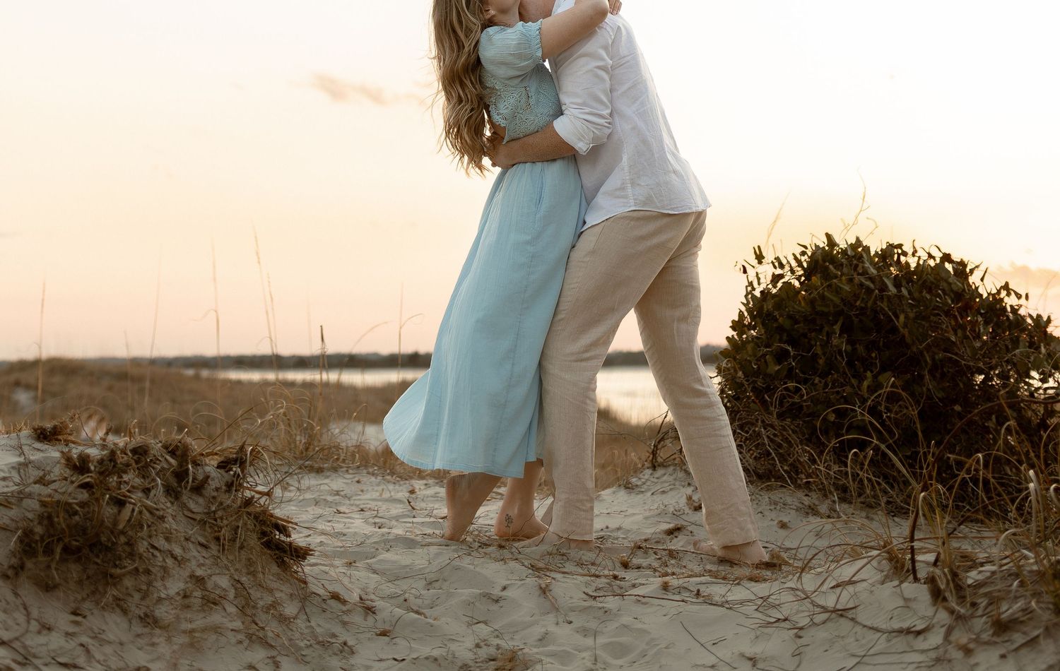 Couple embracing romantically on a sandy beach at sunset, woman in teal dress and man in white shirt.
