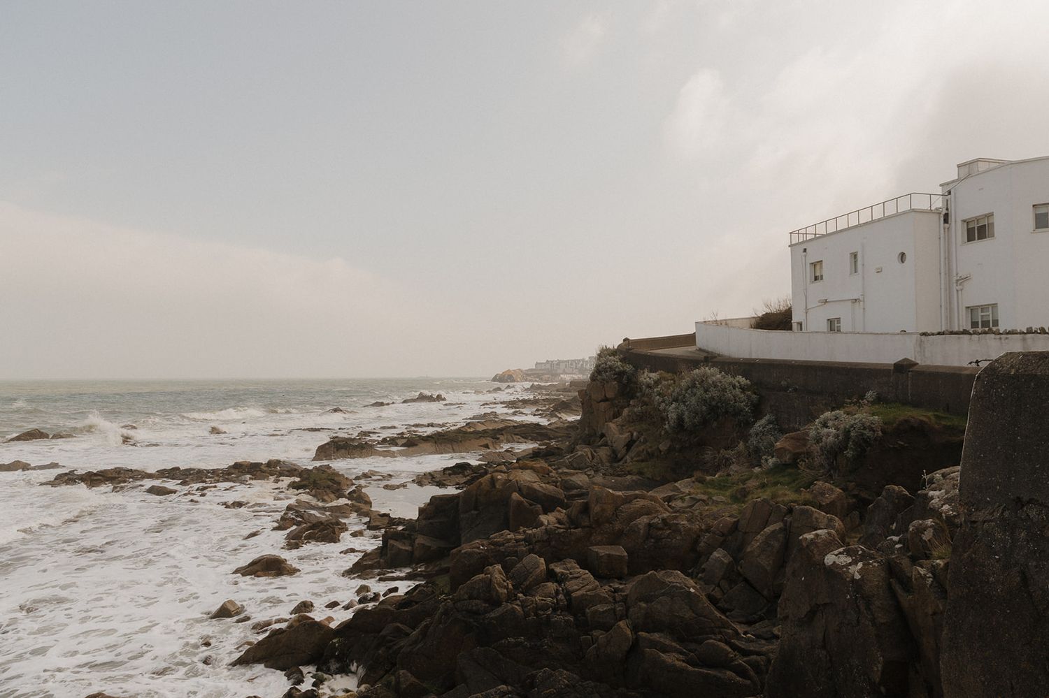 Rocky coastal cliffside with white building overlooking turbulent ocean waves under overcast skies.