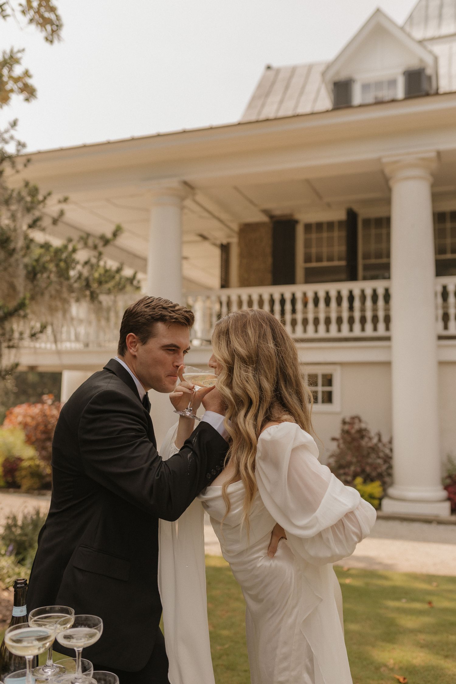 A groom in a black suit kisses his bride's hand outdoors, with a white colonial mansion in the background.