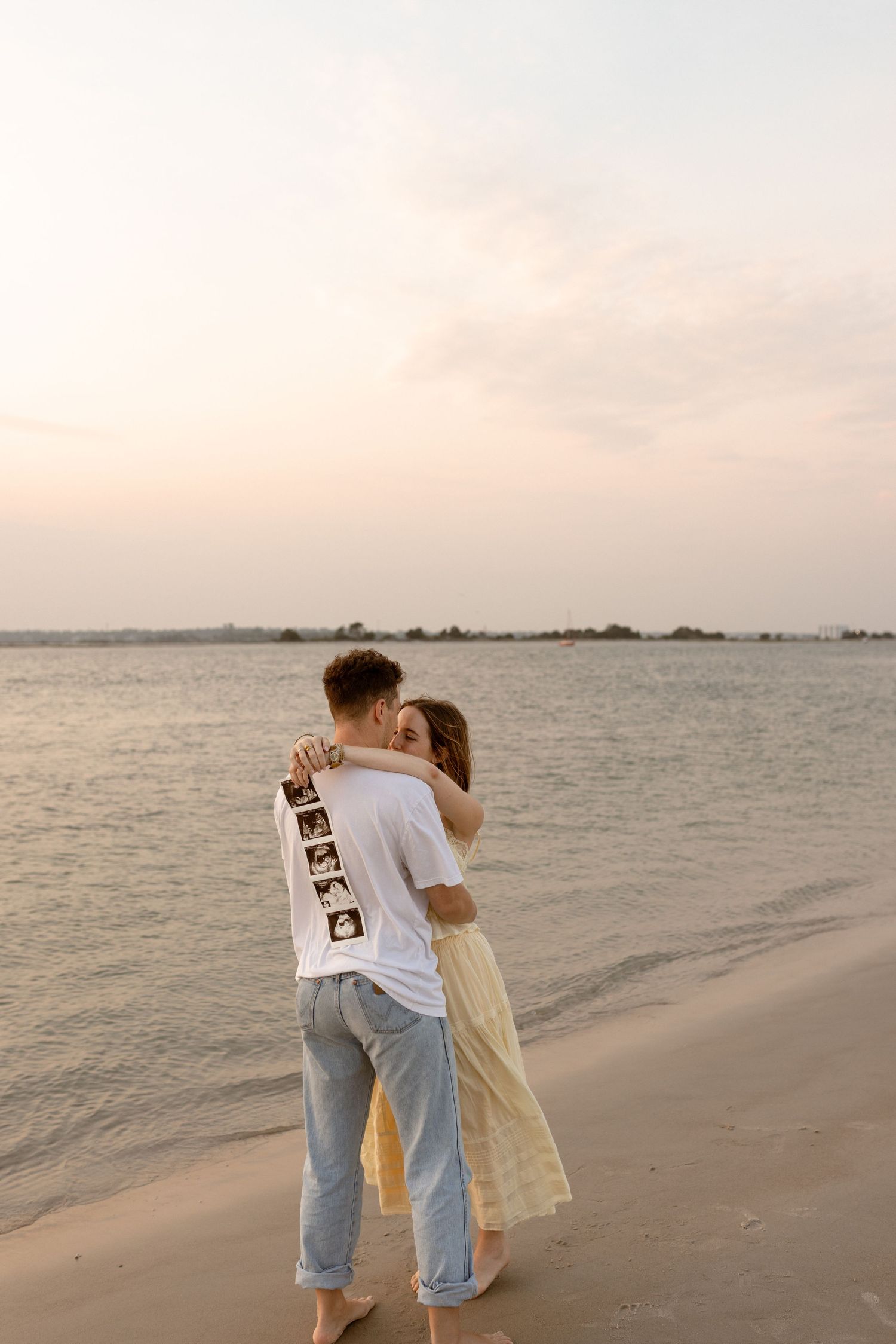 Couple embraces on beach at sunset, man holding ultrasound photos behind his back near calm water.