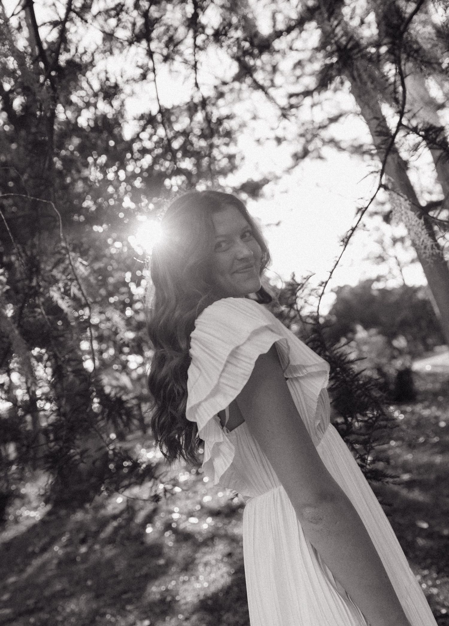 Smiling bride in white ruffled gown glances back in sunlit forest in black and white portrait.