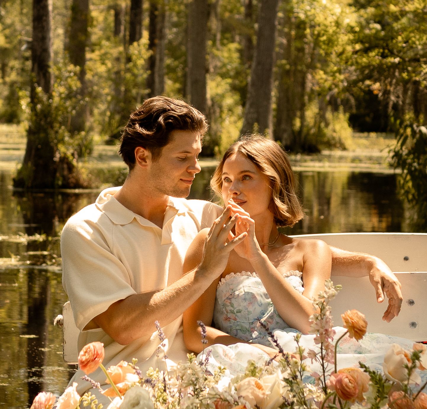 Romantic couple sitting in a boat surrounded by flowers in a sunlit swamp, holding hands tenderly.