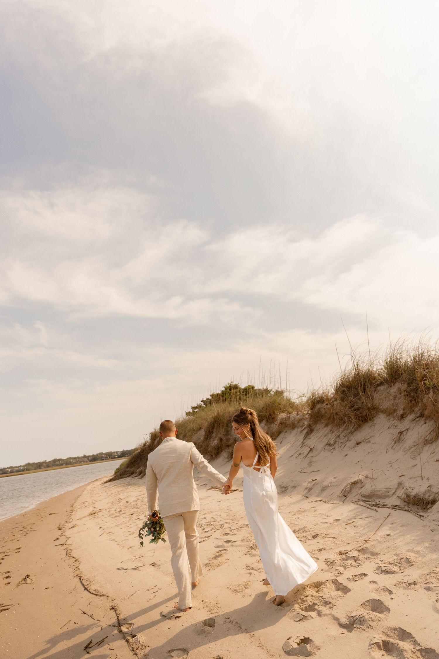 Bride and groom walking hand in hand along a sandy beach with dunes, wearing wedding attire on a sunny day.