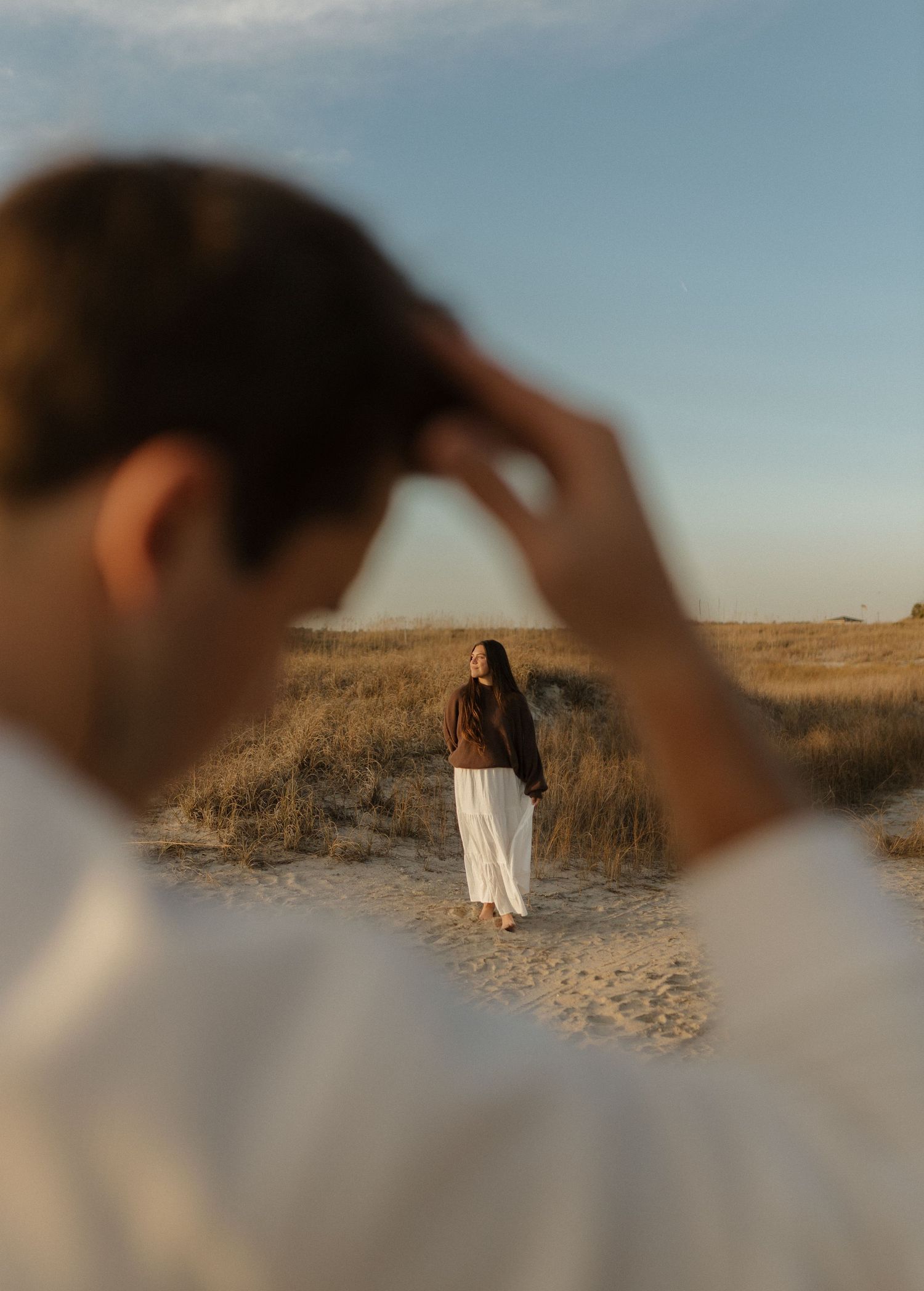 Man in white shirt watches woman in brown top and white skirt standing on sandy beach path.