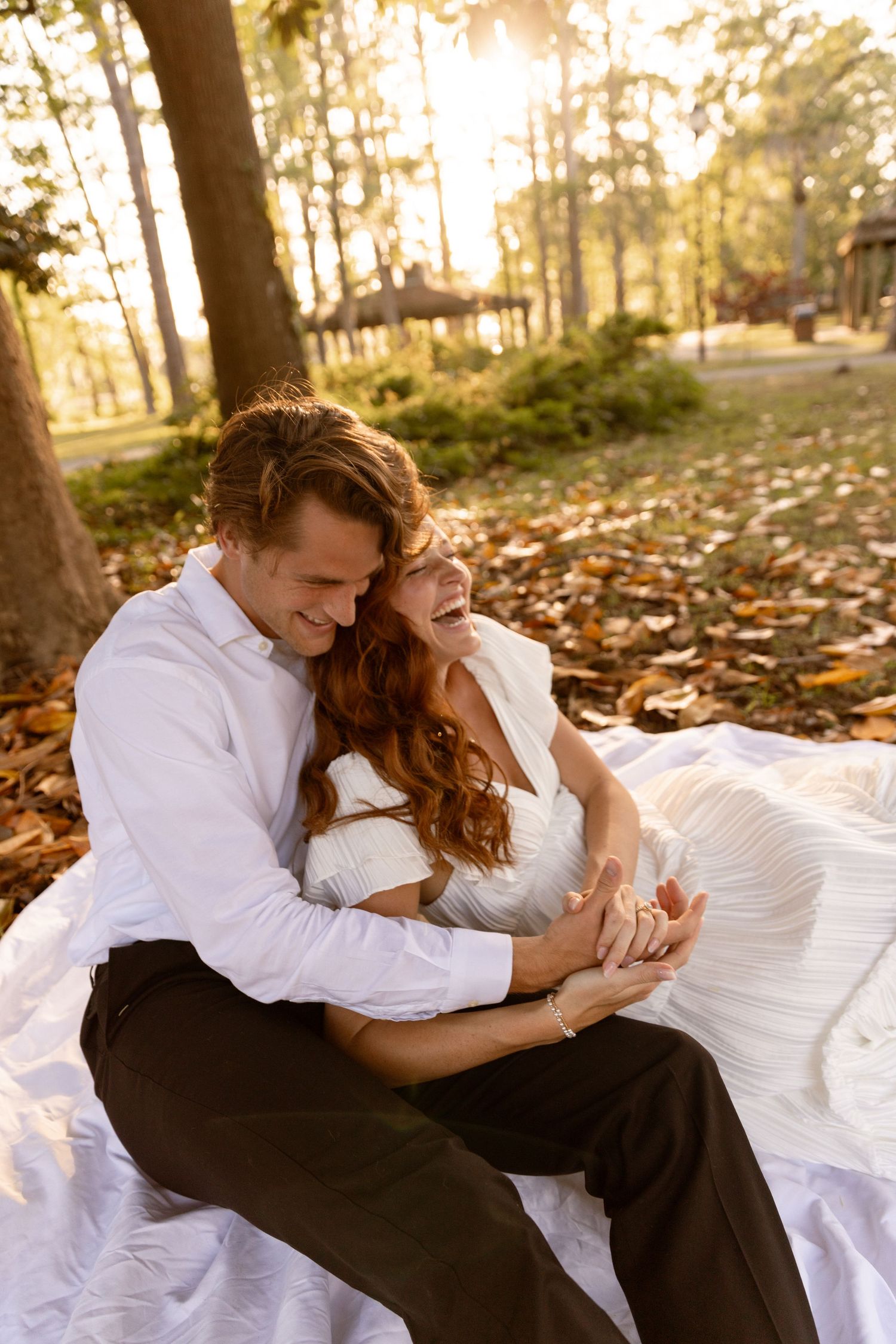 A joyful bride and groom sit together on a white blanket in a sunlit autumn park, sharing a warm embrace.