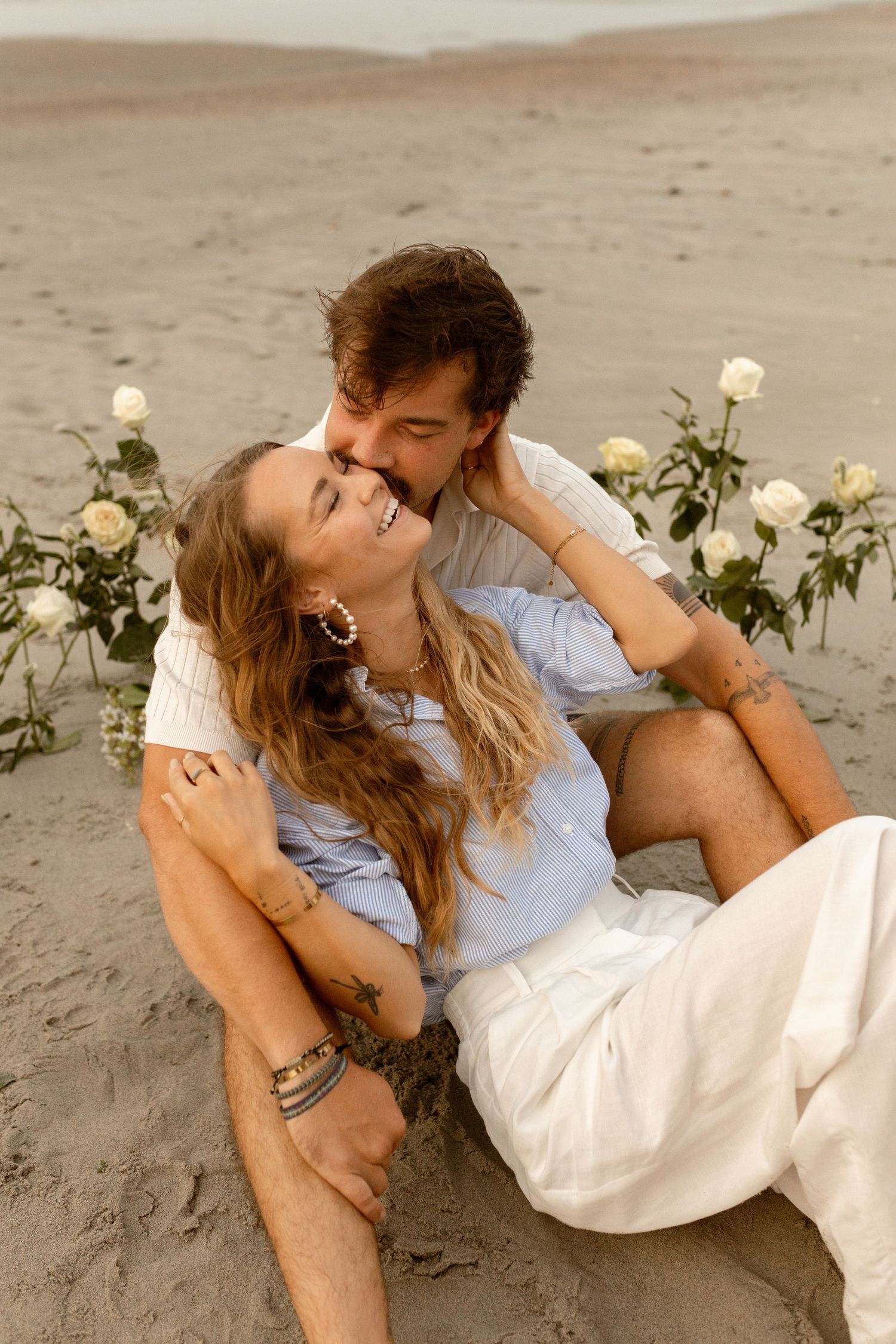 Romantic couple sitting on sandy beach surrounded by white roses, sharing a tender kiss at sunset.
