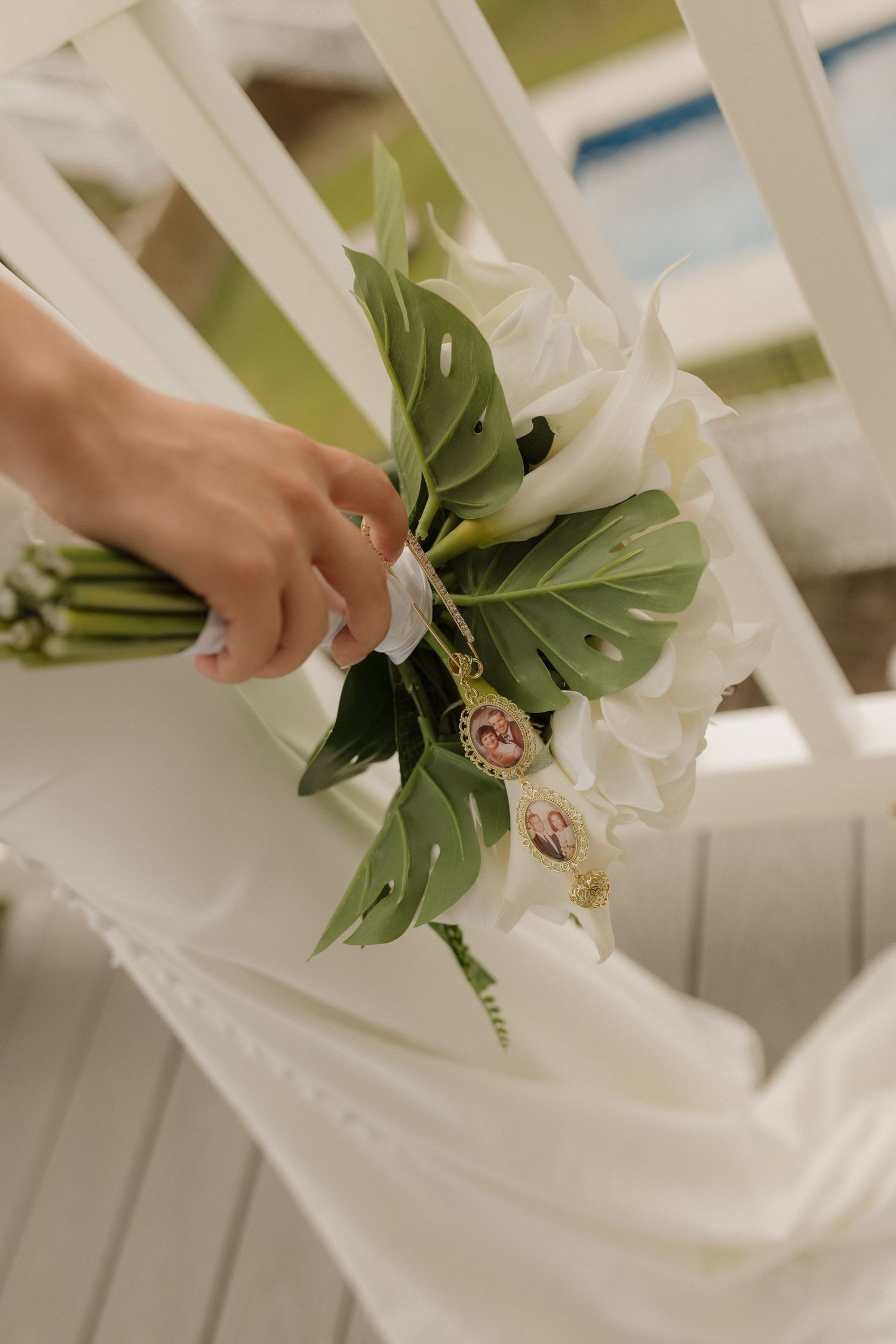A bride holds a elegant white bridal bouquet with monstera leaves and photo charms on a white chair.