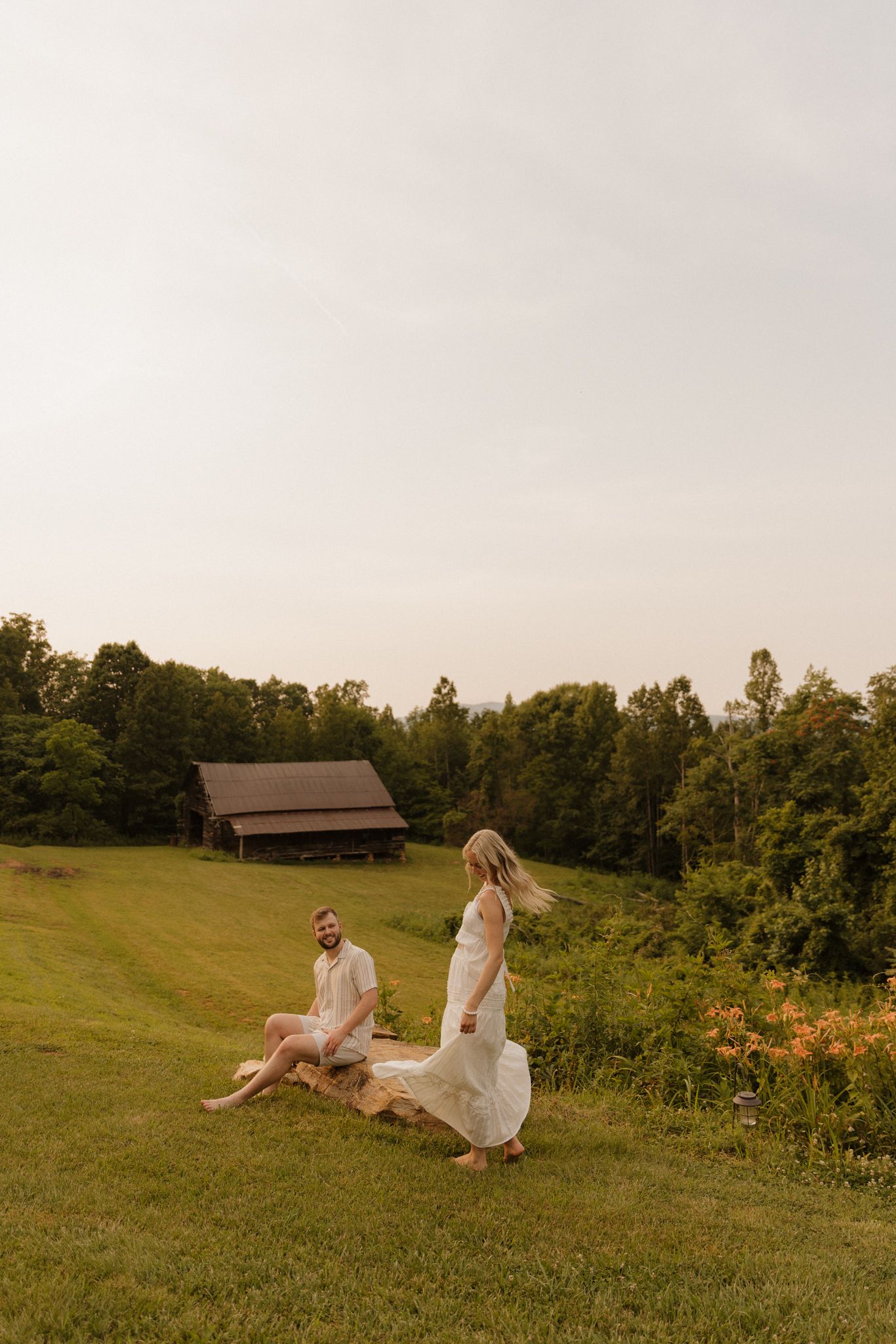 A couple in white attire enjoys a golden hour moment on a lush green farm field with a rustic barn backdrop.