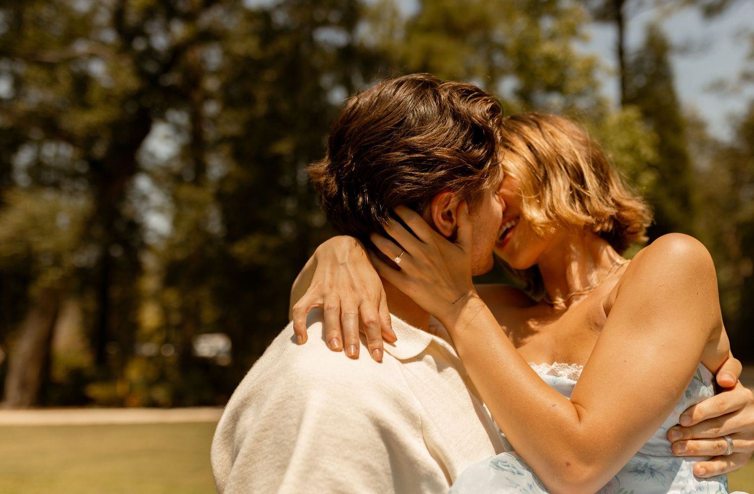 Couple sharing a romantic kiss outdoors surrounded by tall green trees on a sunny day.