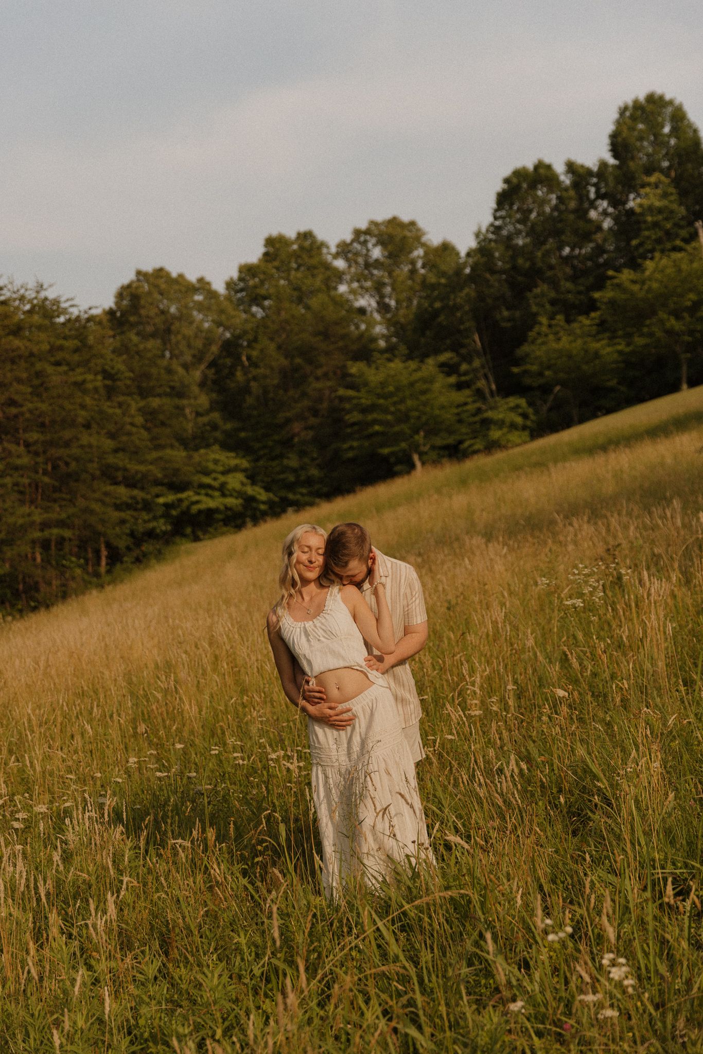 A couple shares a tender embrace in a golden meadow, the woman in a white dress cradling her baby bump.