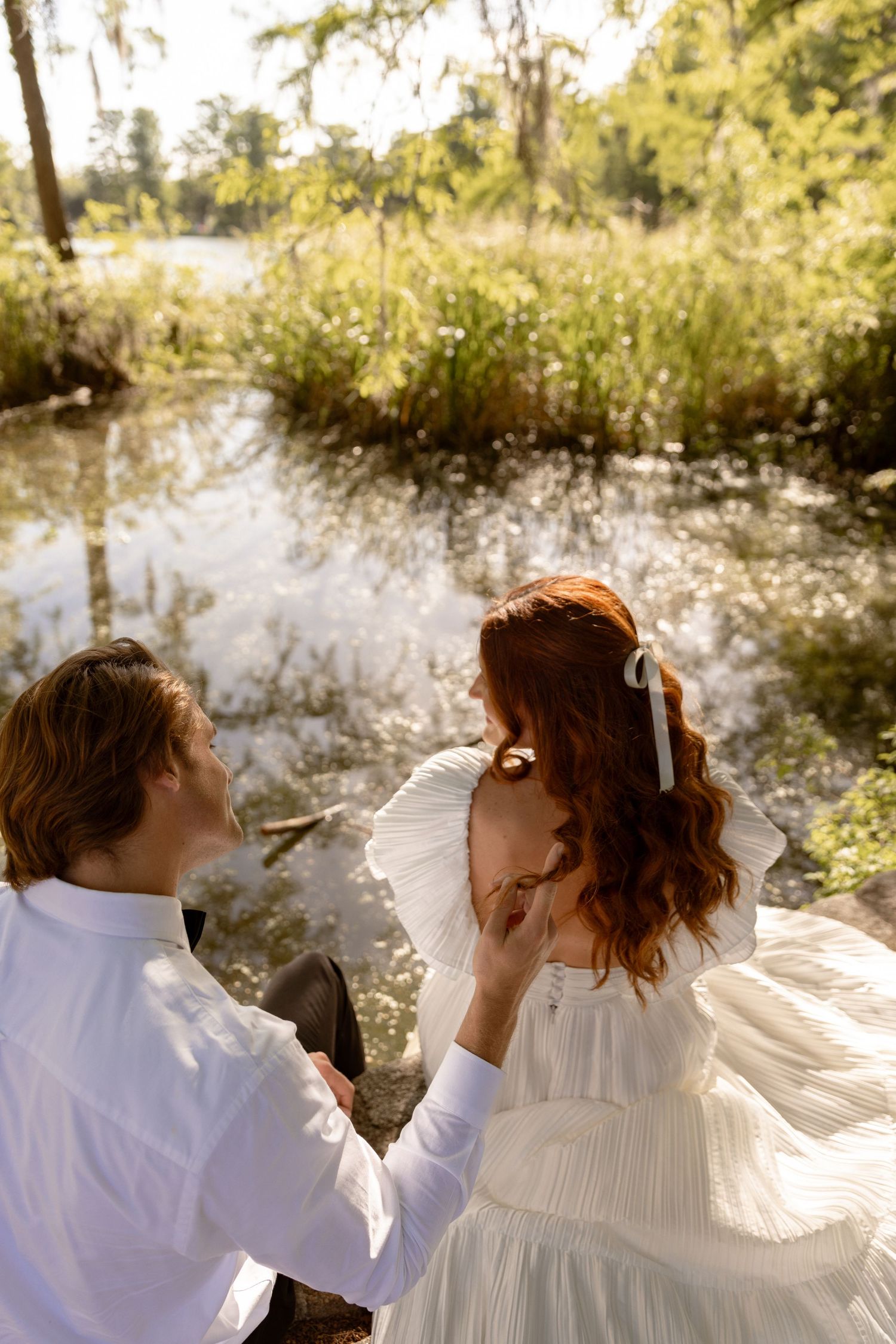 A couple in white attire sits romantically by a sunlit pond surrounded by lush greenery.