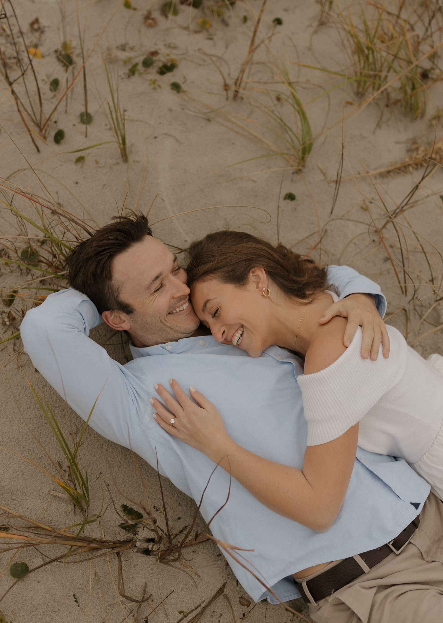 Smiling couple lies together on sandy dunes, woman hugging man in light blue shirt outdoors.