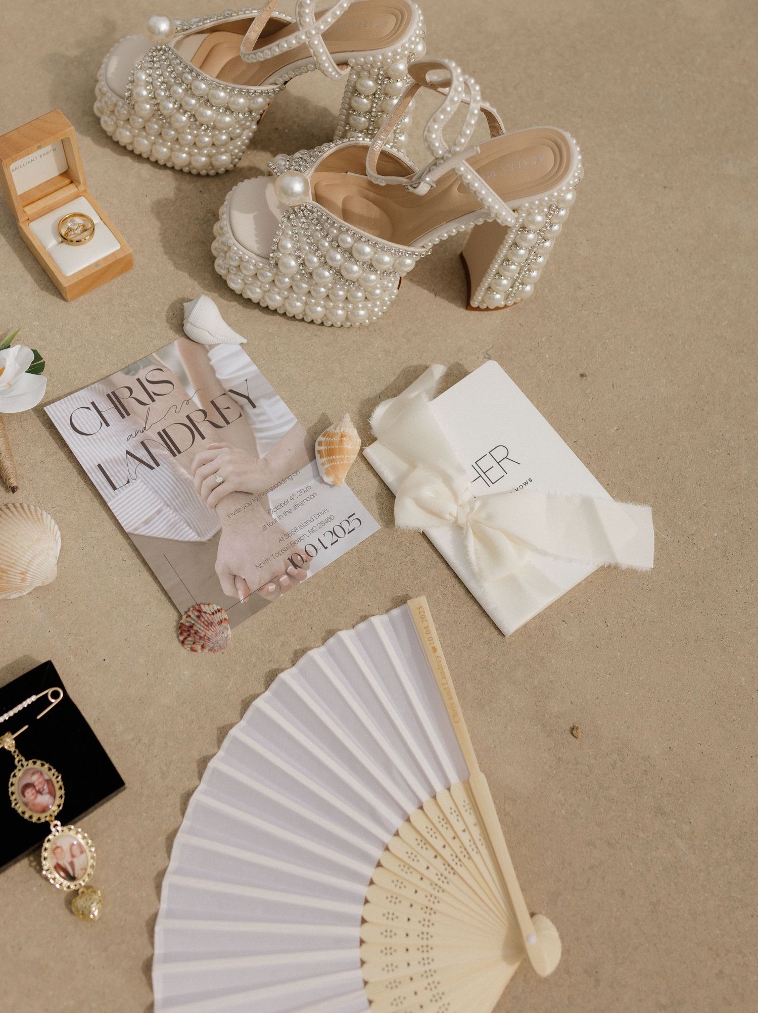 Pearl-embellished bridal heels, hand fan, rings, and accessories flatlay on beige sandy surface.