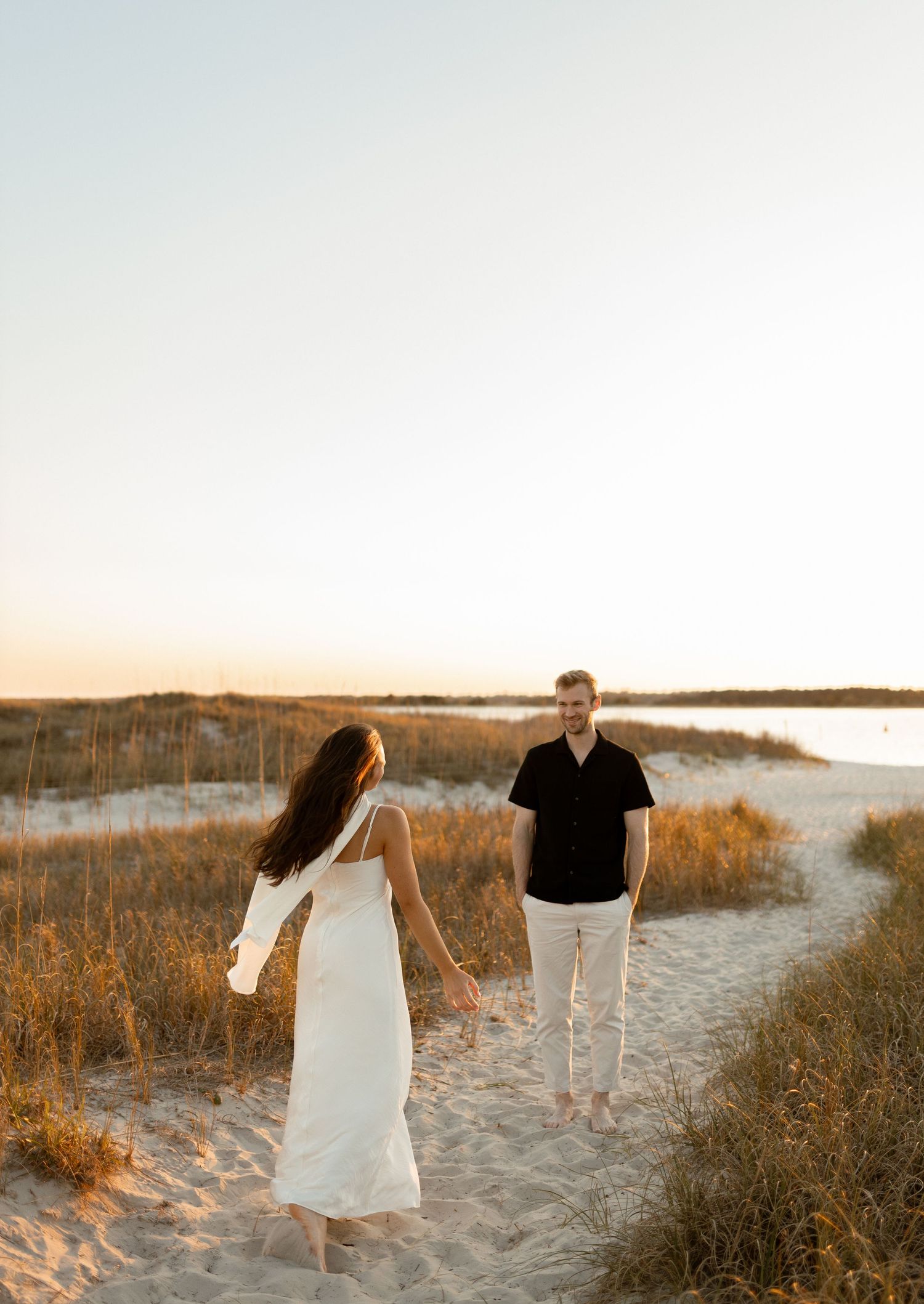 Couple holding hands on sandy beach path at golden hour, woman in white dress, man in black shirt.
