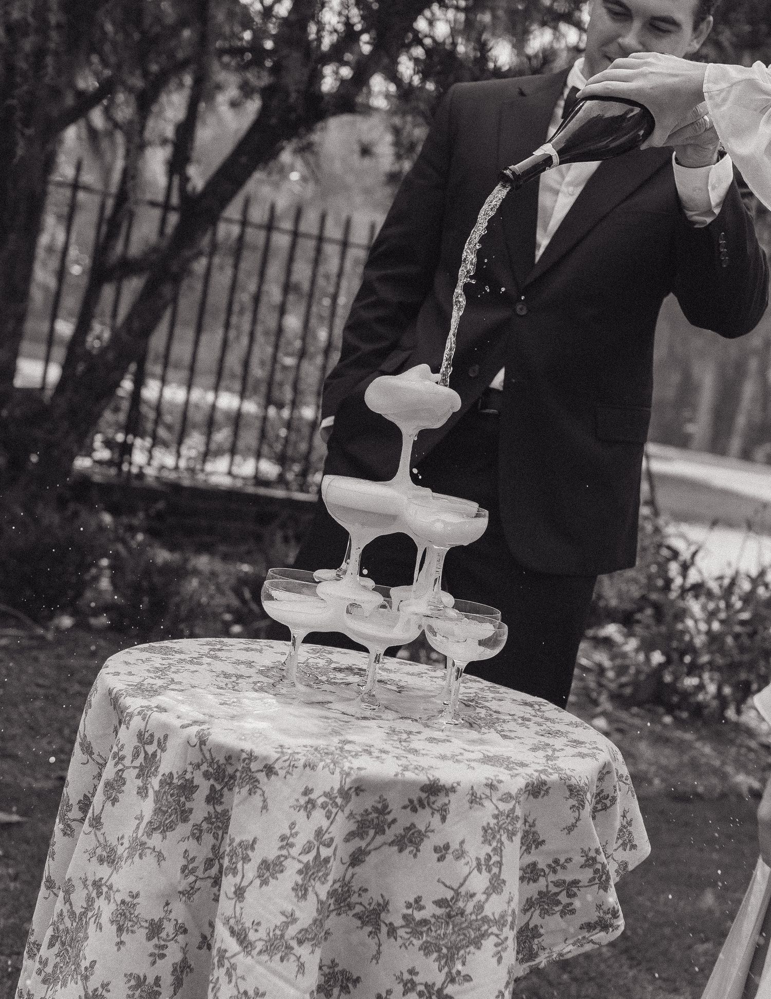 Man in black suit pouring champagne into a tower of glasses at an outdoor event.