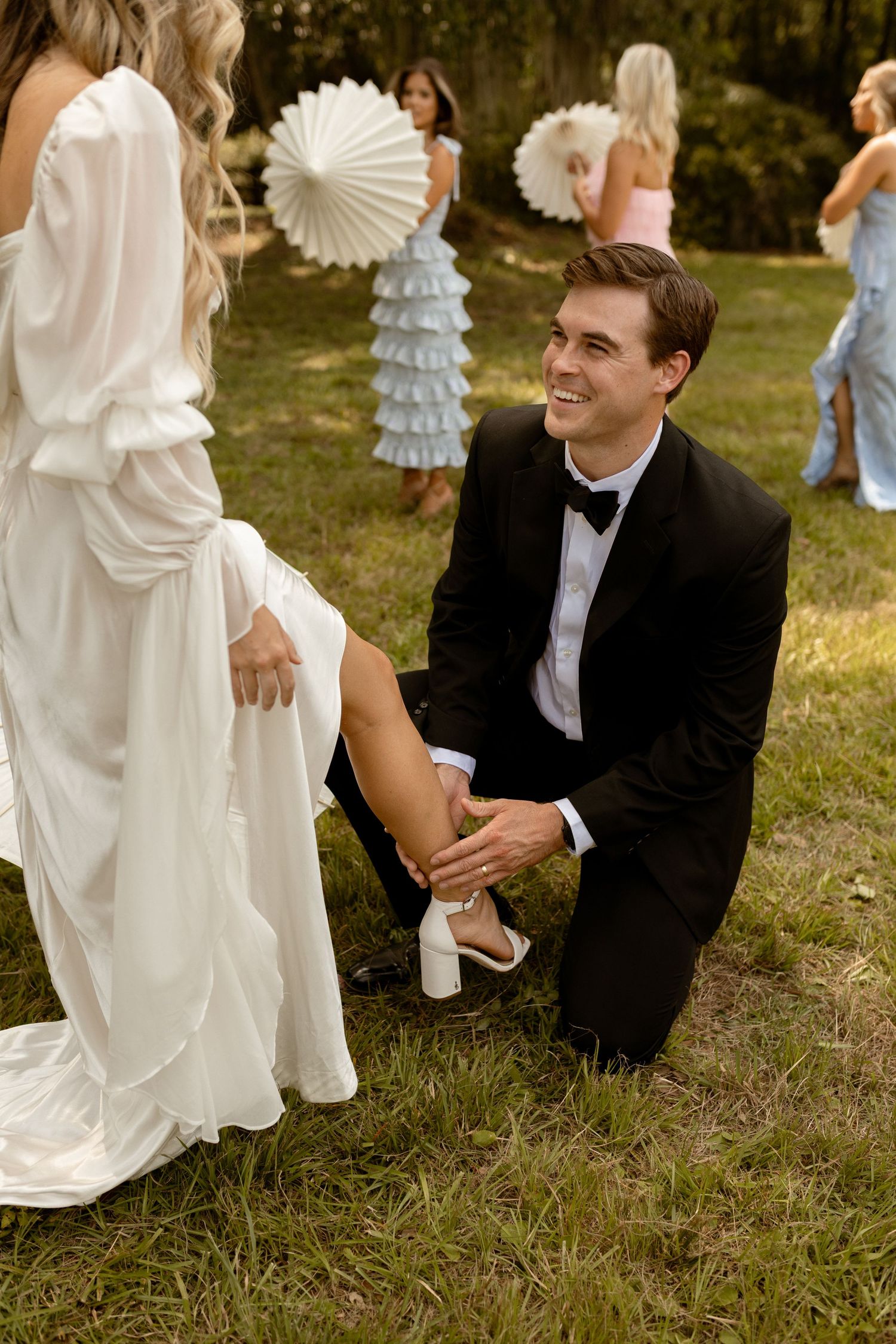 Groom kneeling on grass, smiling while adjusting bride's shoe at outdoor wedding ceremony.
