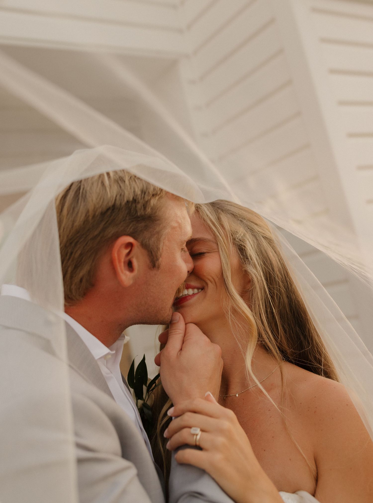 Bride and groom kiss under flowing white veil with white architectural ceiling in background.