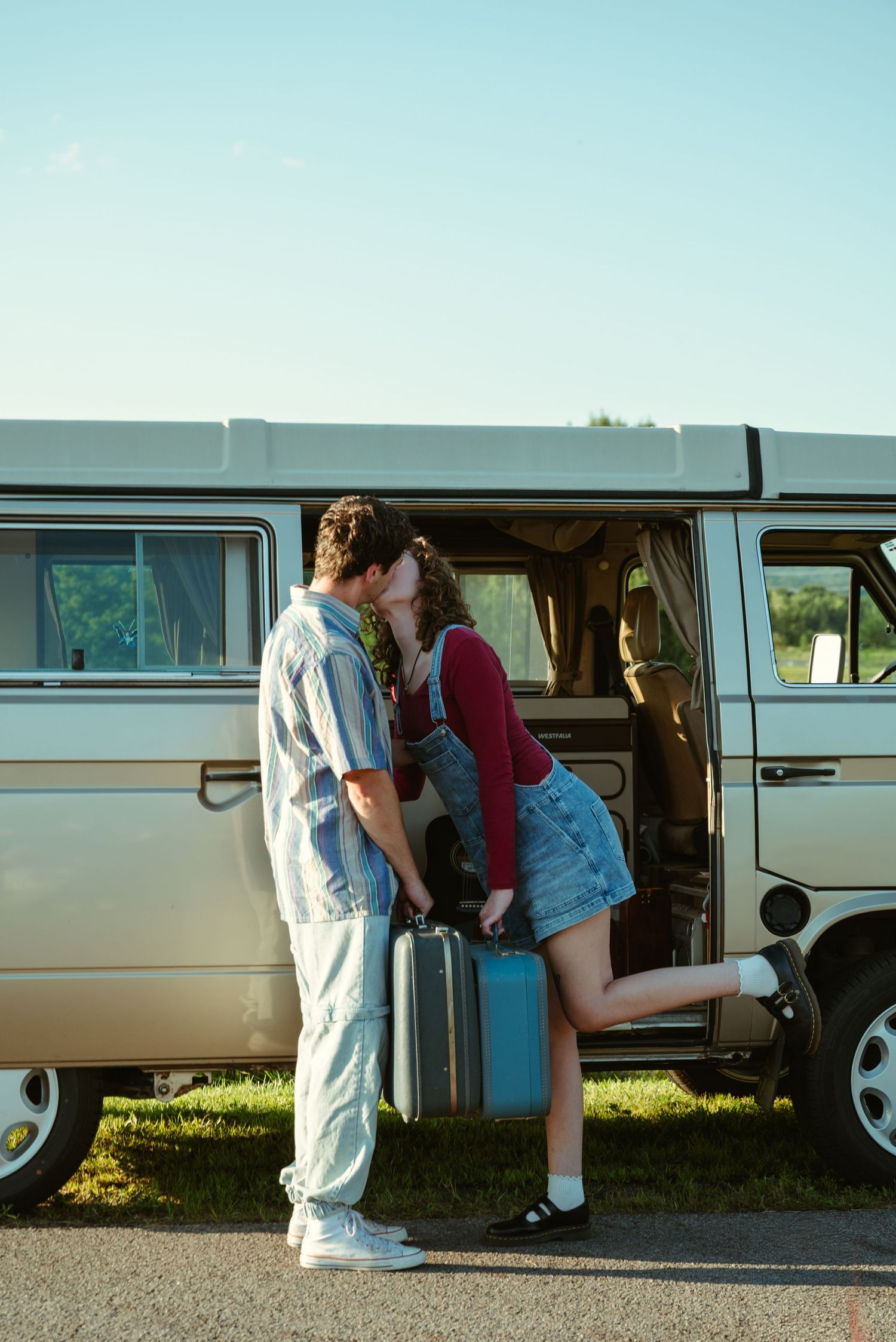 A couple shares a romantic kiss beside a vintage van, with the woman lifting her leg while holding a suitcase.