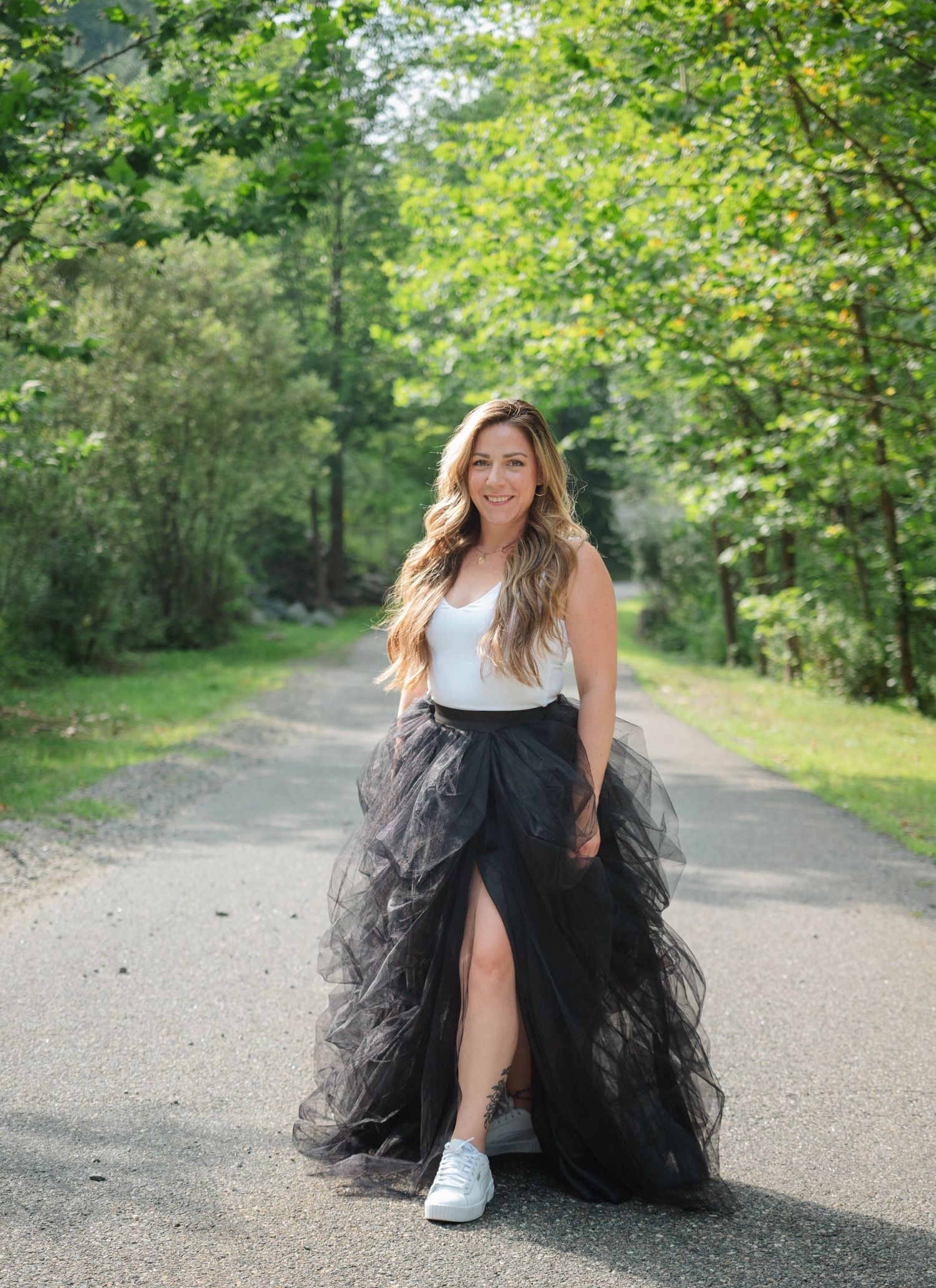 Woman in black tulle skirt and white top walks confidently down tree-lined path wearing sneakers.