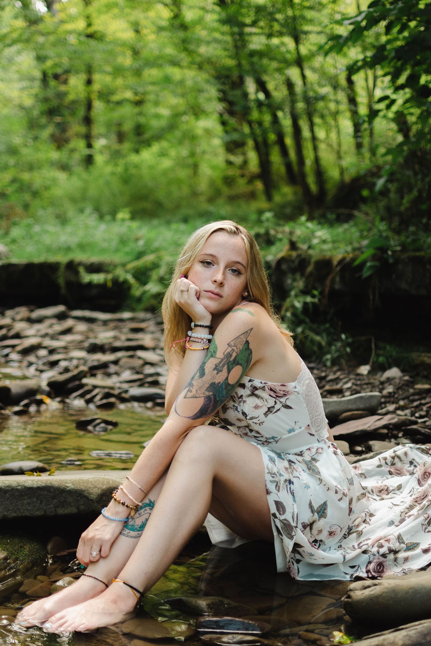 Tattooed blonde woman in floral dress sits barefoot on rocks beside forest stream.