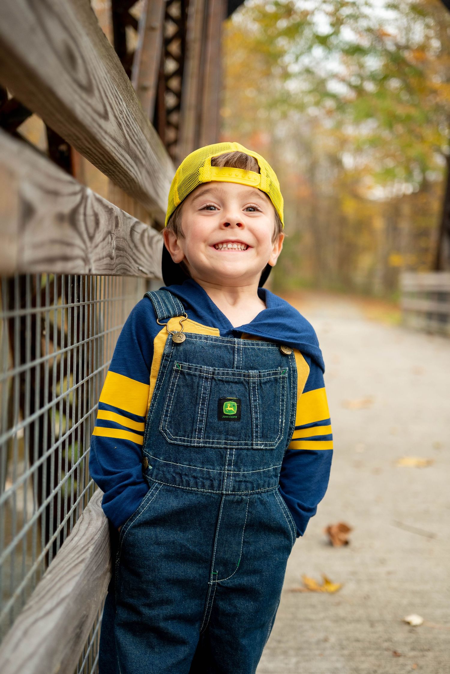 Happy young boy in John Deere denim overalls and yellow cap looking up outdoors in autumn.