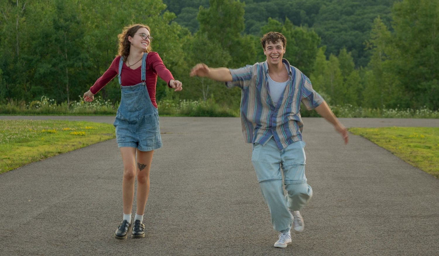 Two young people joyfully running hand-in-hand down a scenic road surrounded by lush green trees.