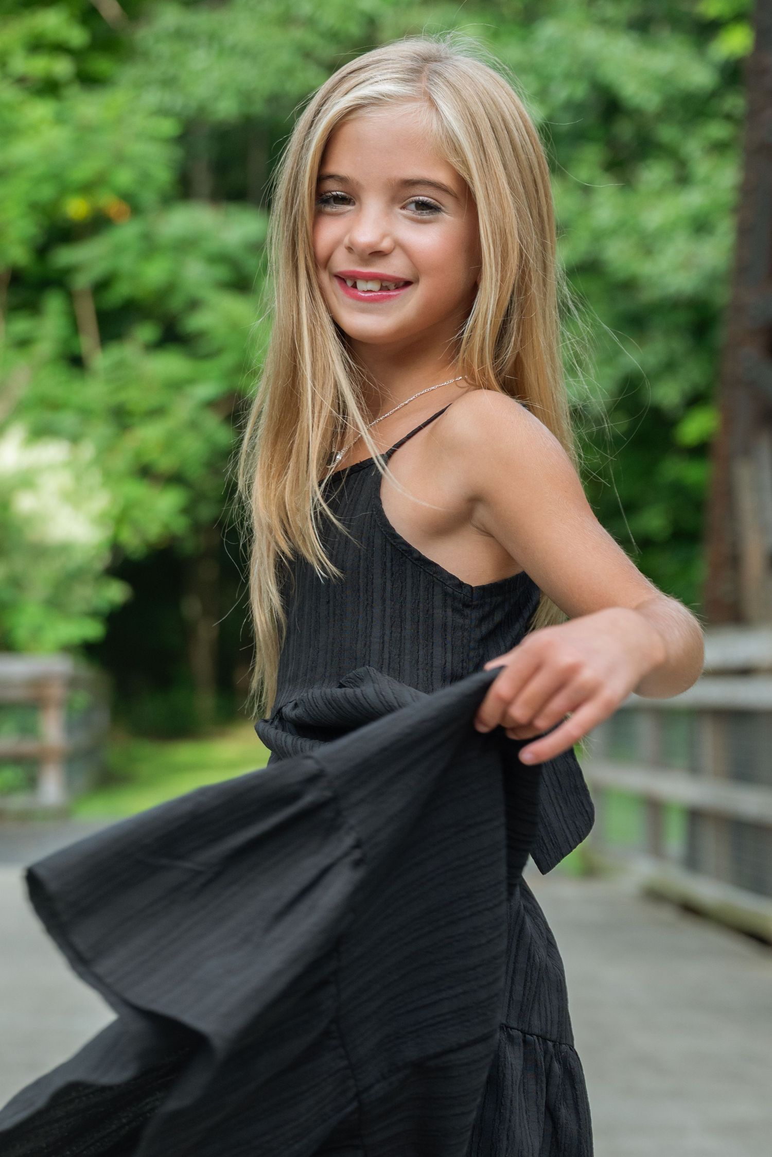 Young blonde girl in black dress twirling outdoors on wooden bridge, smiling joyfully with green trees background.