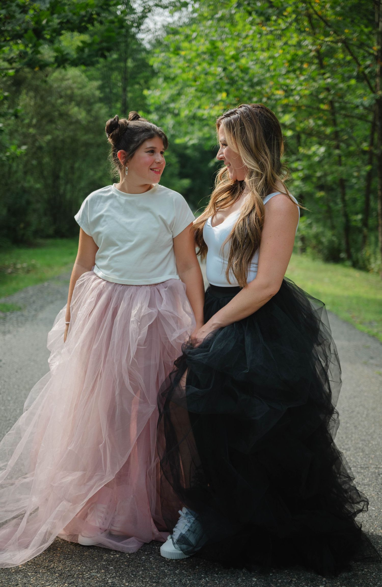 Two women in white tops and tulle skirts, pink and black, holding hands on a tree-lined path.