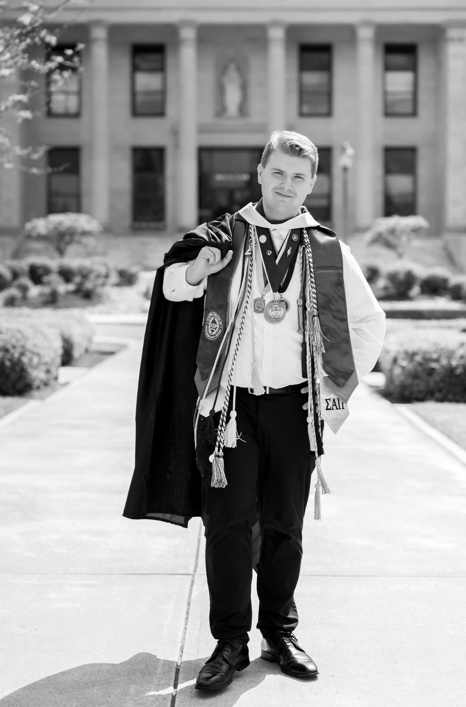 Graduate in academic regalia with medals and sash poses proudly in front of a classic university building.