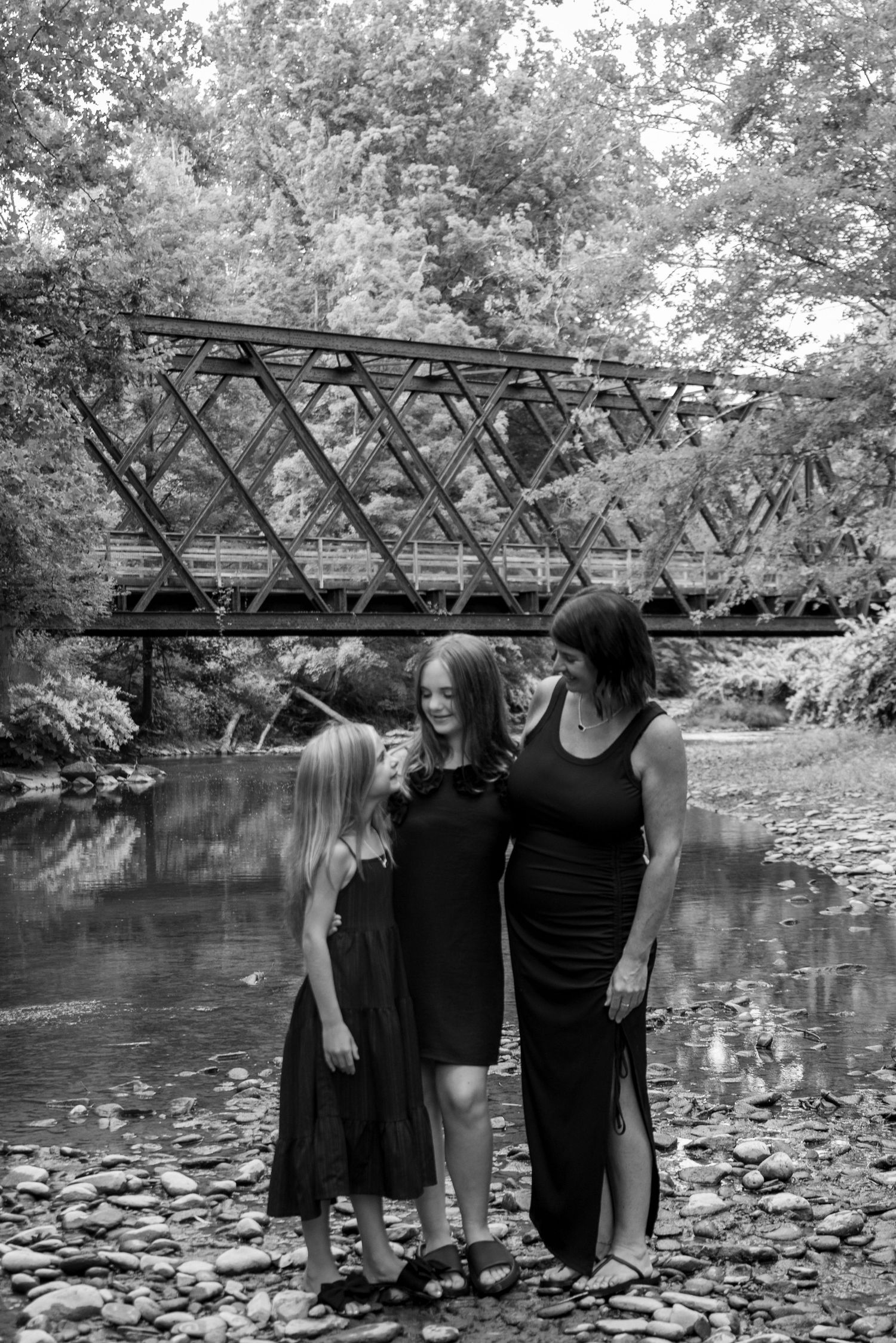 Three women in black dresses smile together on a rocky riverbank with a steel truss bridge and trees in the background.