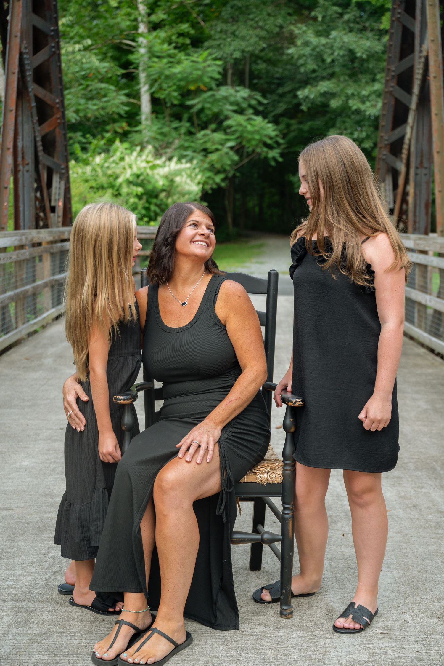 A woman in a black dress sits on a chair on a bridge, smiling at two girls in black outfits outdoors.