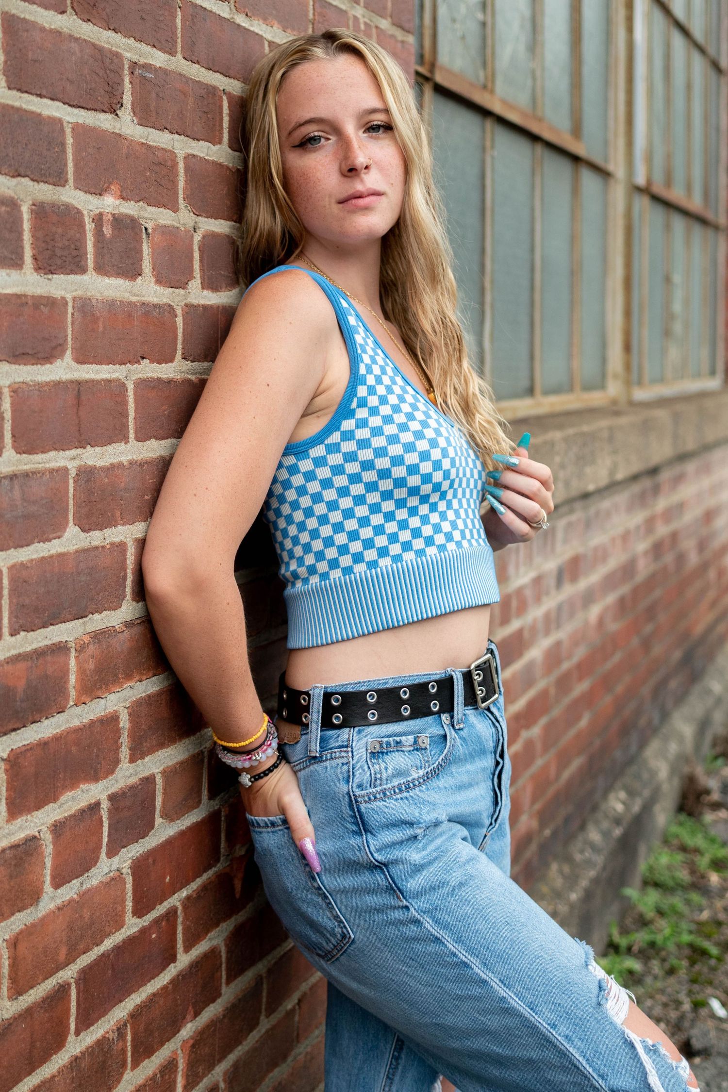 Young blonde woman in blue checkered crop top and jeans leaning against a brick wall.