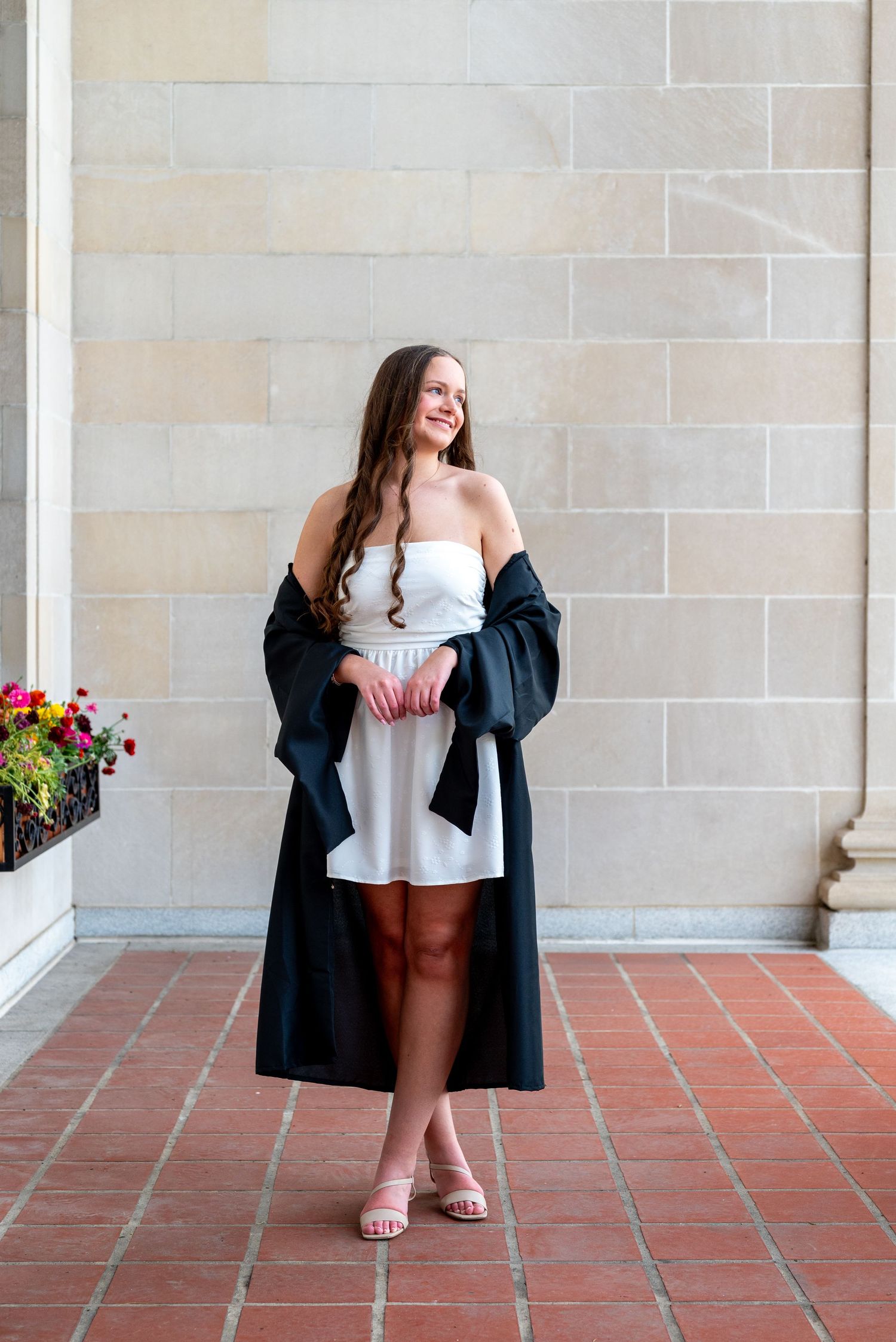 Smiling graduate in white dress with black graduation gown poses against stone wall in outdoor corridor.