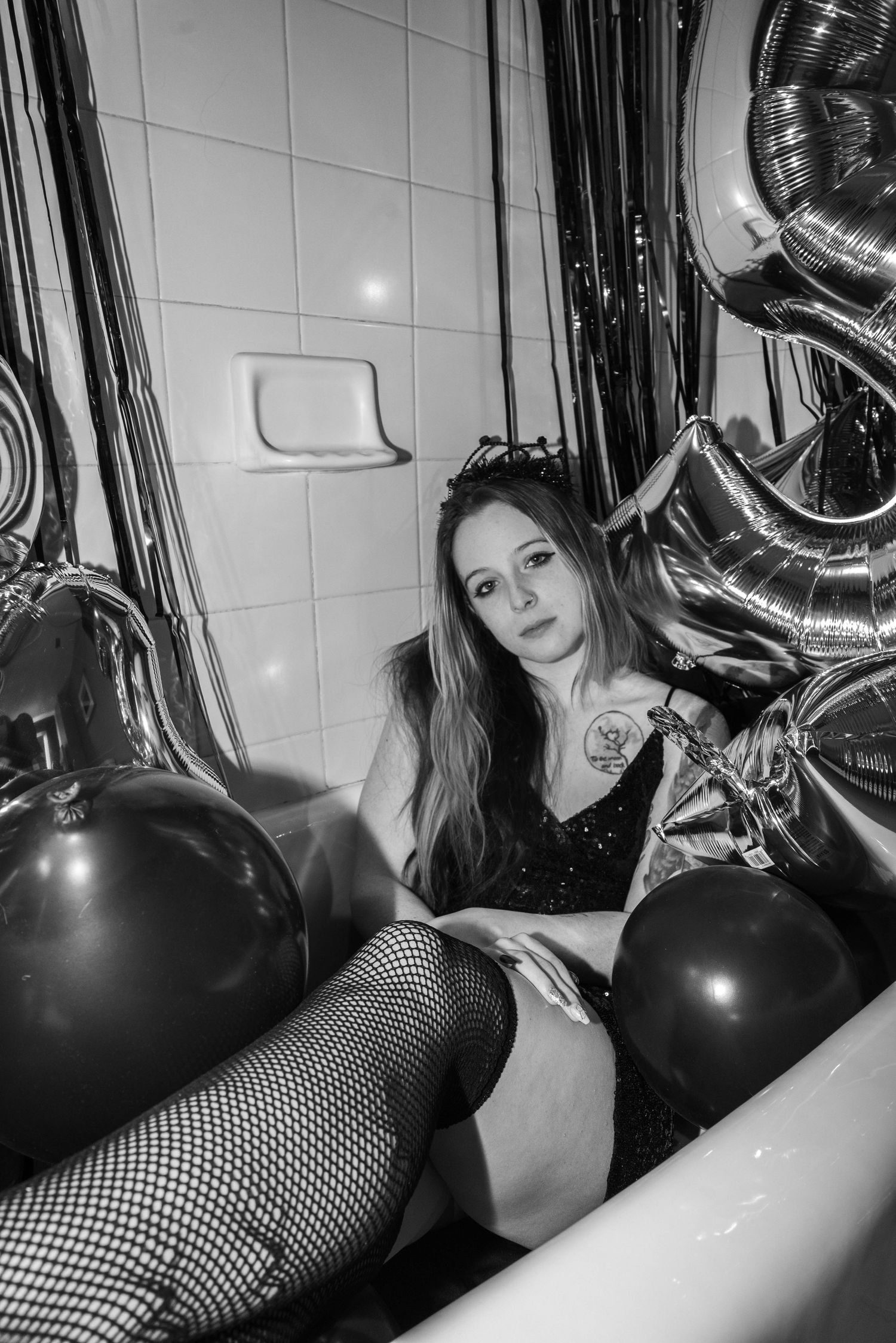 Black and white photo of woman in fishnet stockings sitting in bathtub surrounded by balloons.