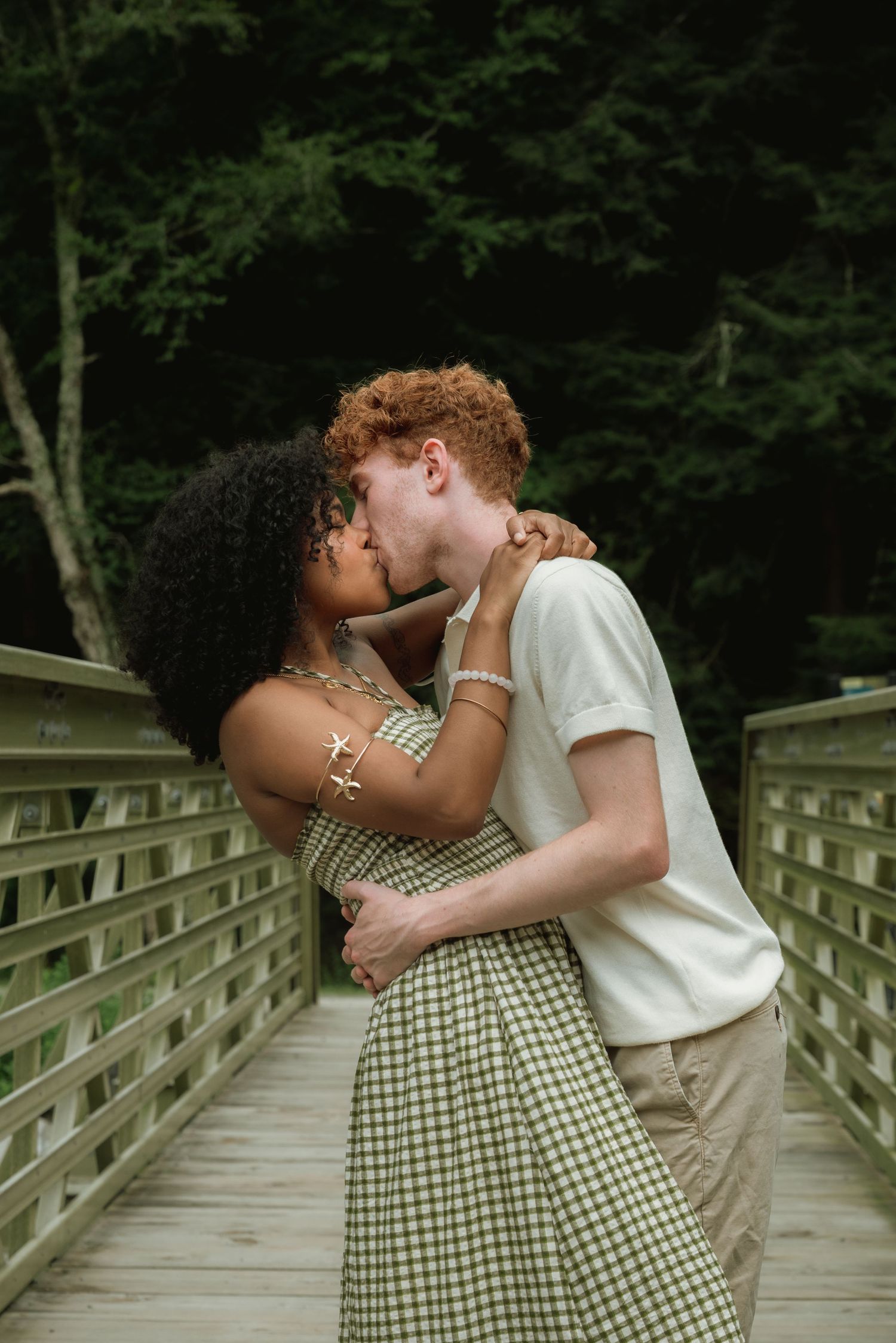 A young interracial couple shares a romantic kiss on a wooden bridge surrounded by lush green trees.
